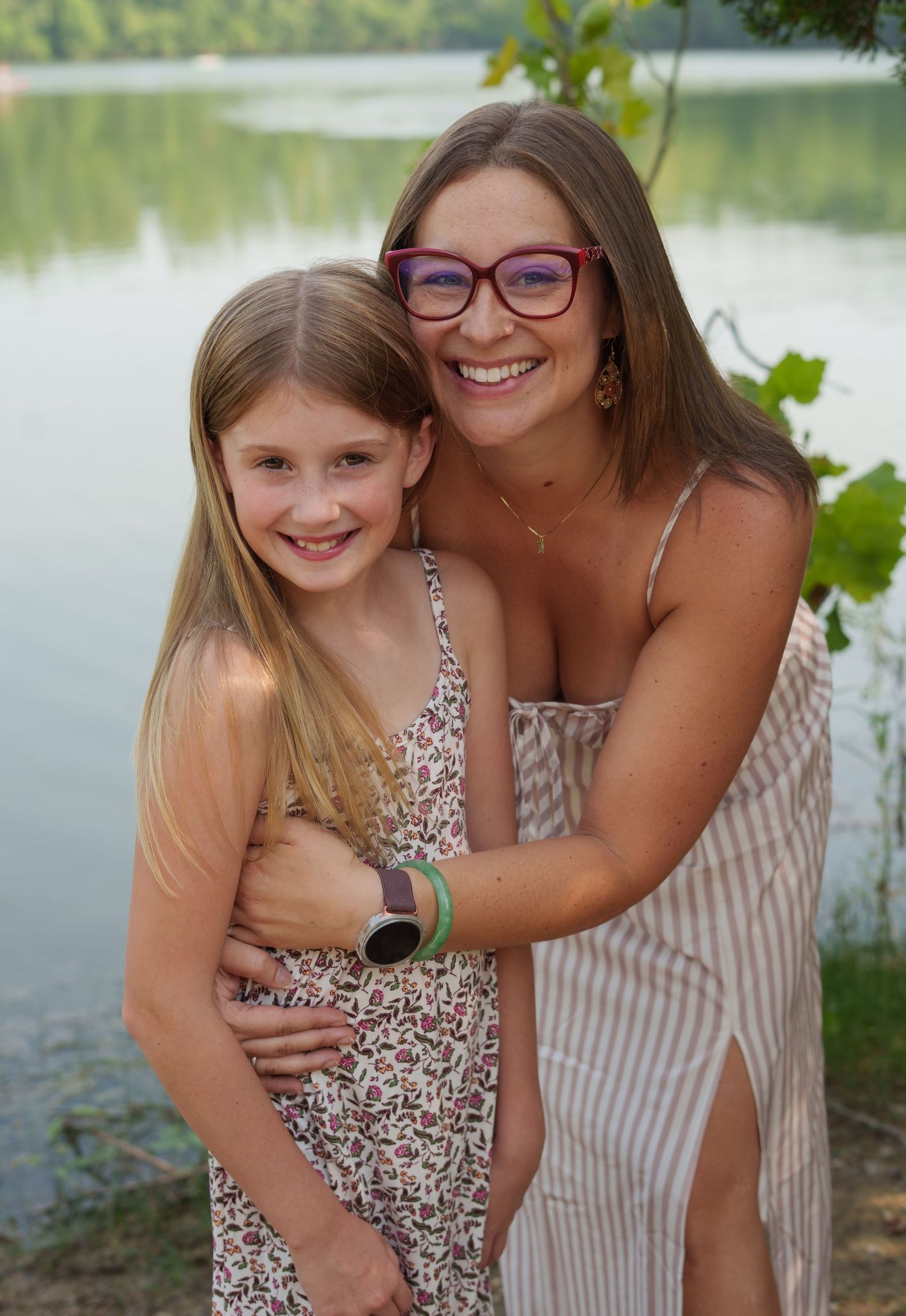 A woman with red glasses hugs a young blonde girl by a lake, both smiling in summer dresses.