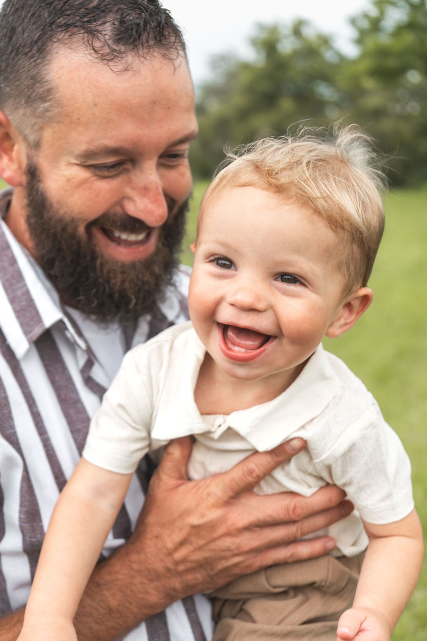 A joyful outdoor portrait shows genuine laughter and tender bonding between a bearded adult and happy toddler.
