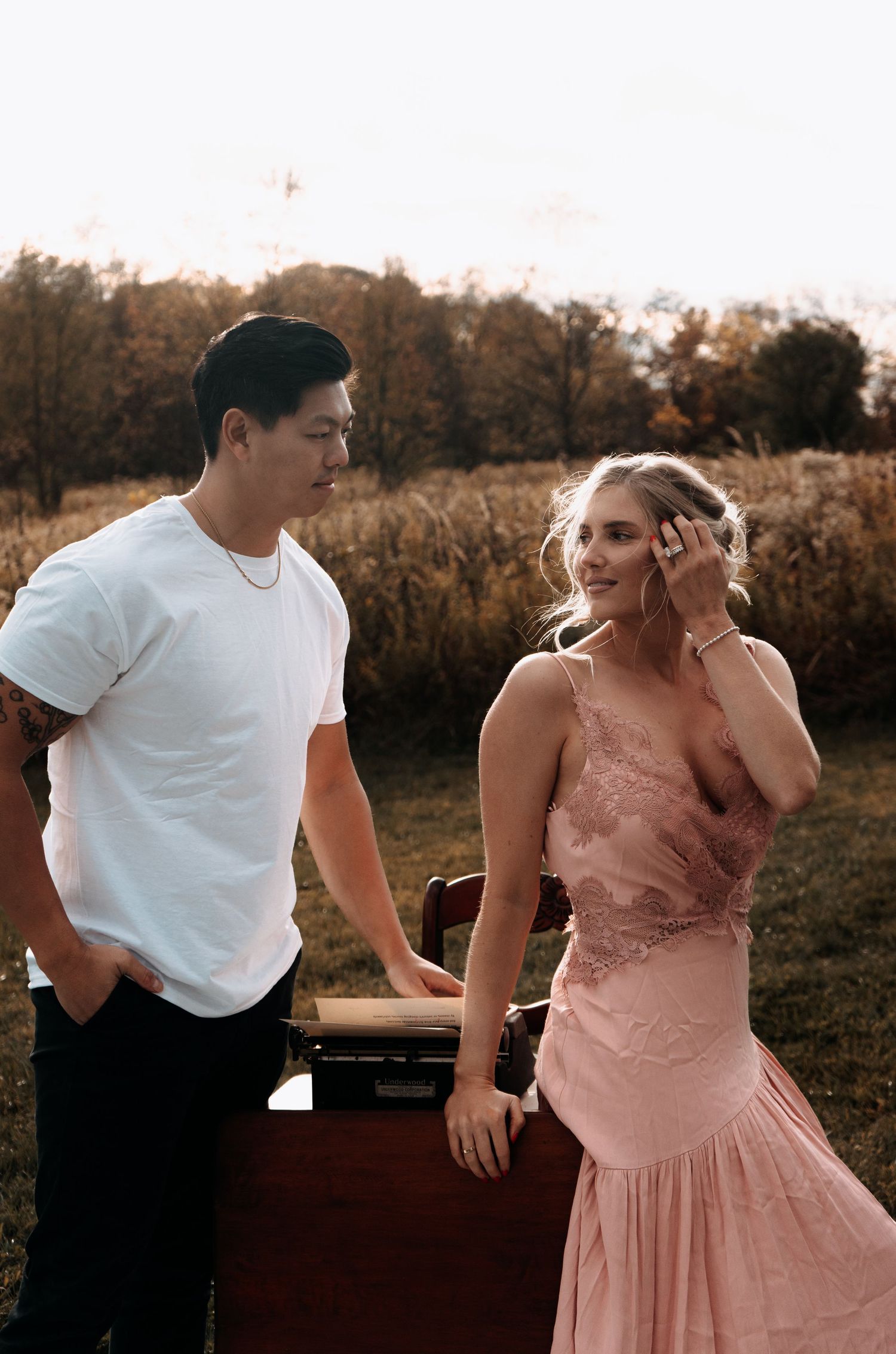 A couple in pink dress and white shirt share romantic moments at sunset in a rustic outdoor setting.
