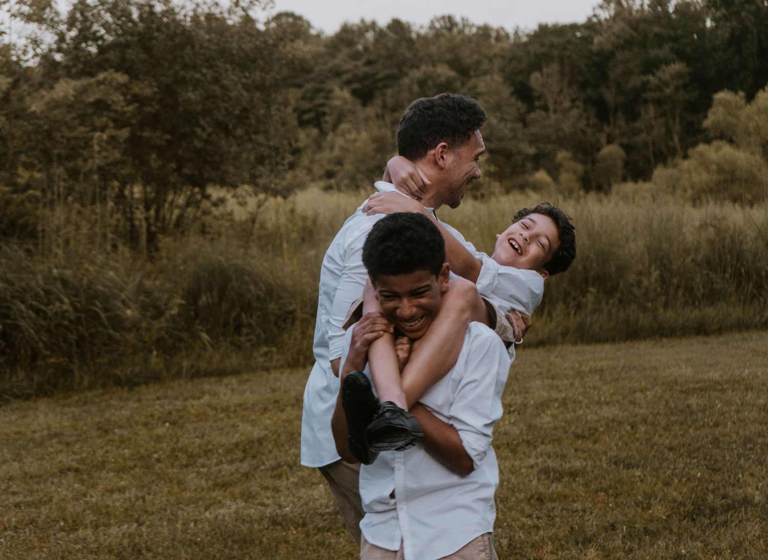 Family wearing white clothes embrace in a candid moment while standing in a grassy field at sunset.