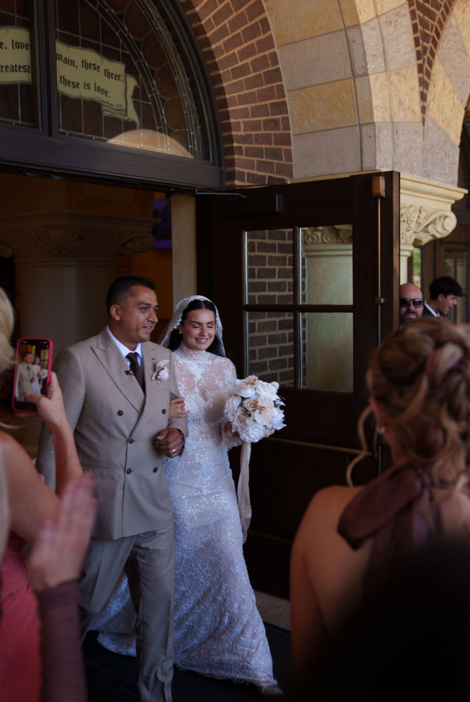 Bride and groom exit church in wedding attire, guests photograph the joyful couple after ceremony.