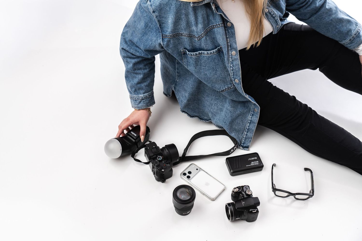 Photographer sitting on white floor with DSLR camera, lens, flash, smartphone, and glasses arranged nearby.