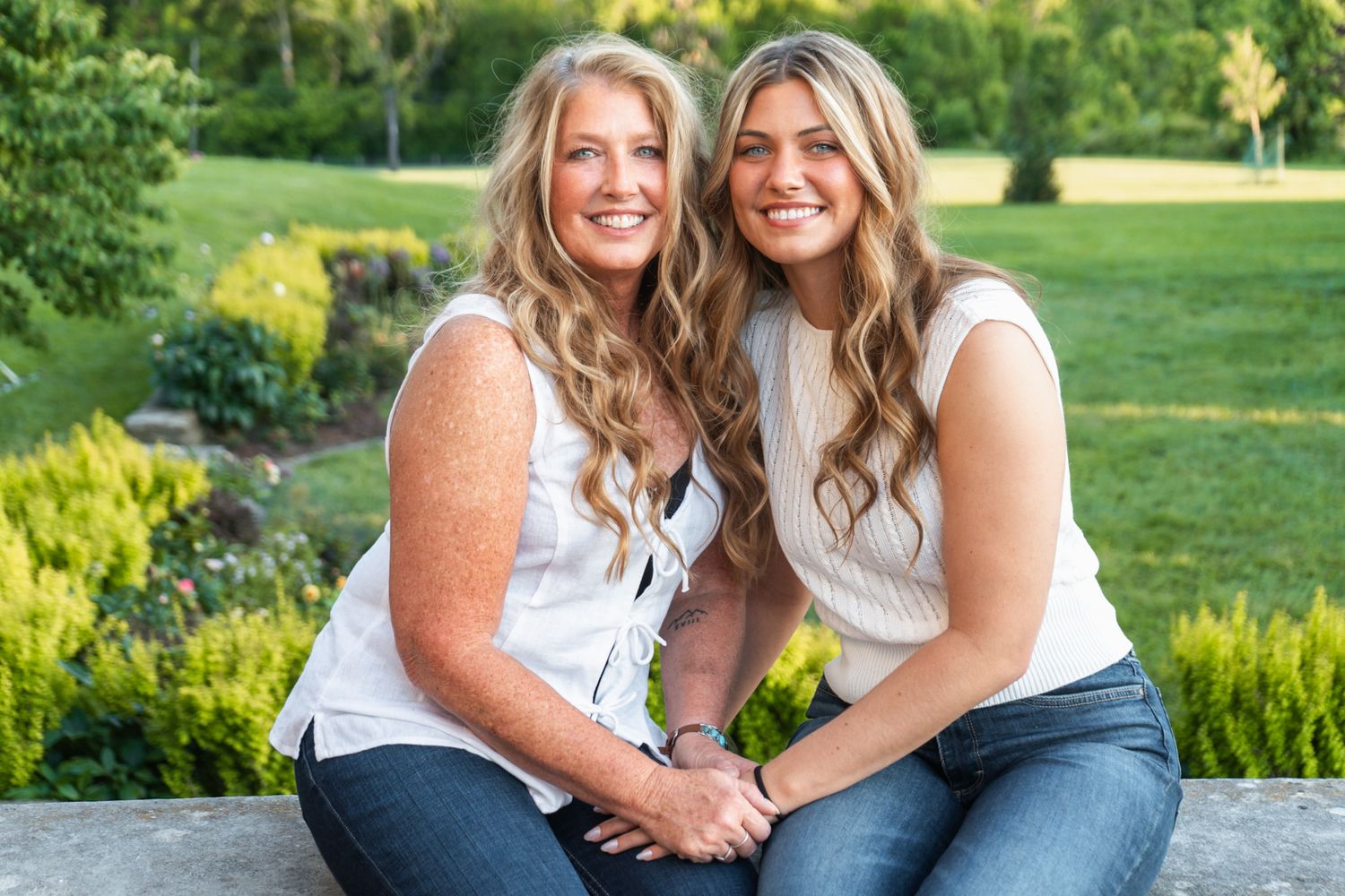 Two women in white tops and jeans sitting together outdoors in a garden setting.