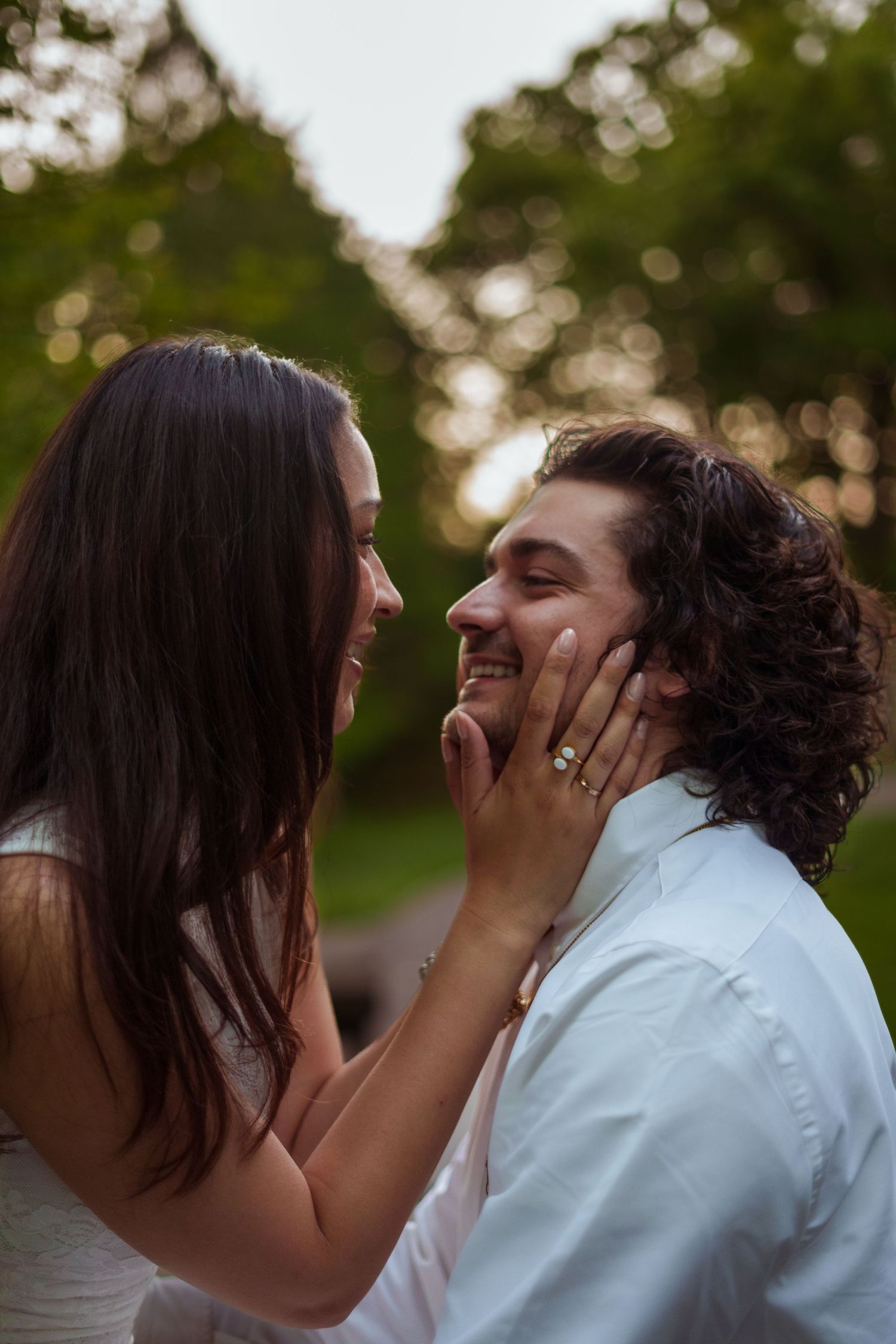 Couple laughing together outdoors, woman holding man's face showing engagement ring, green trees in background.