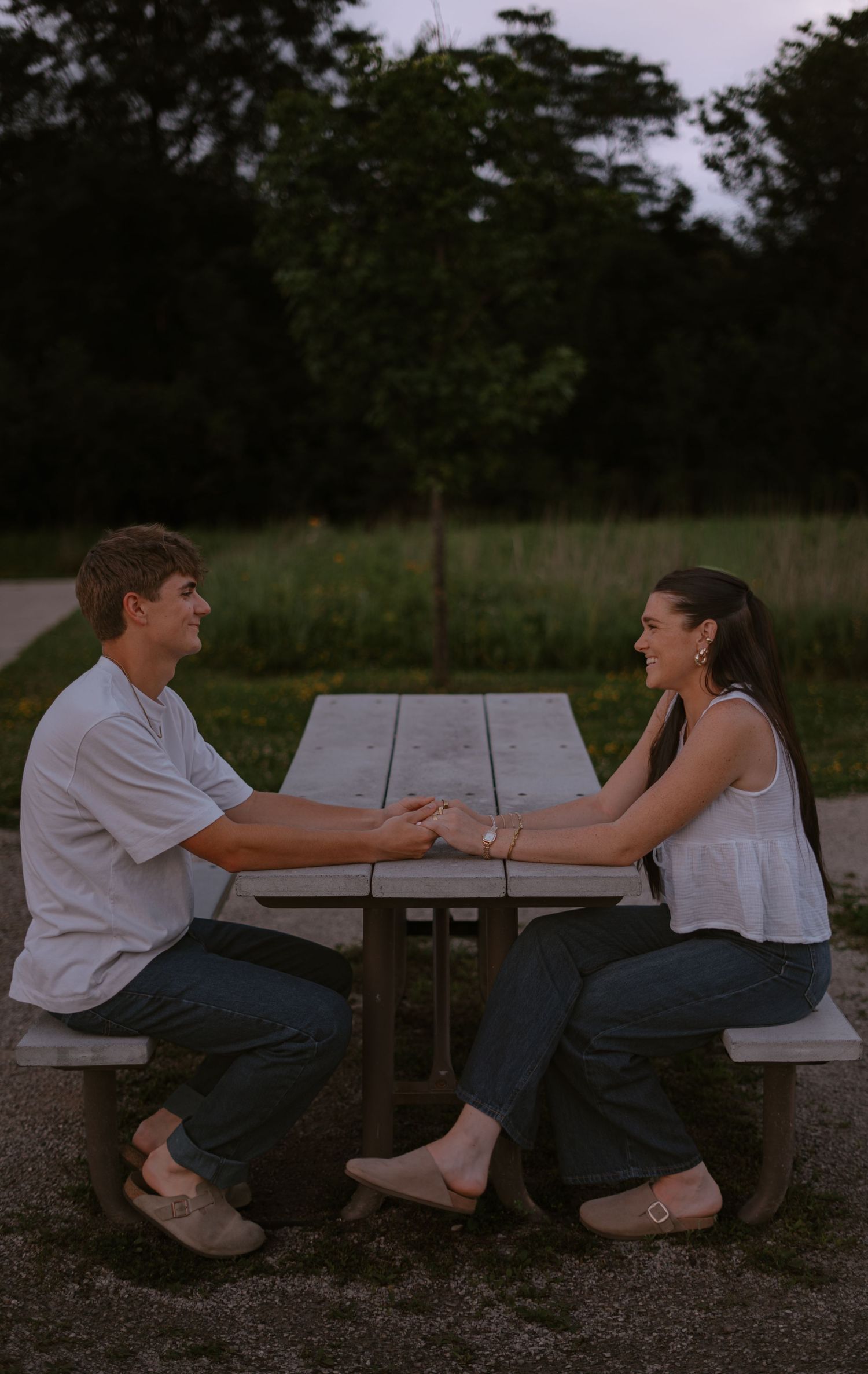 A young couple holds hands across a picnic table outdoors at dusk, smiling at each other romantically.