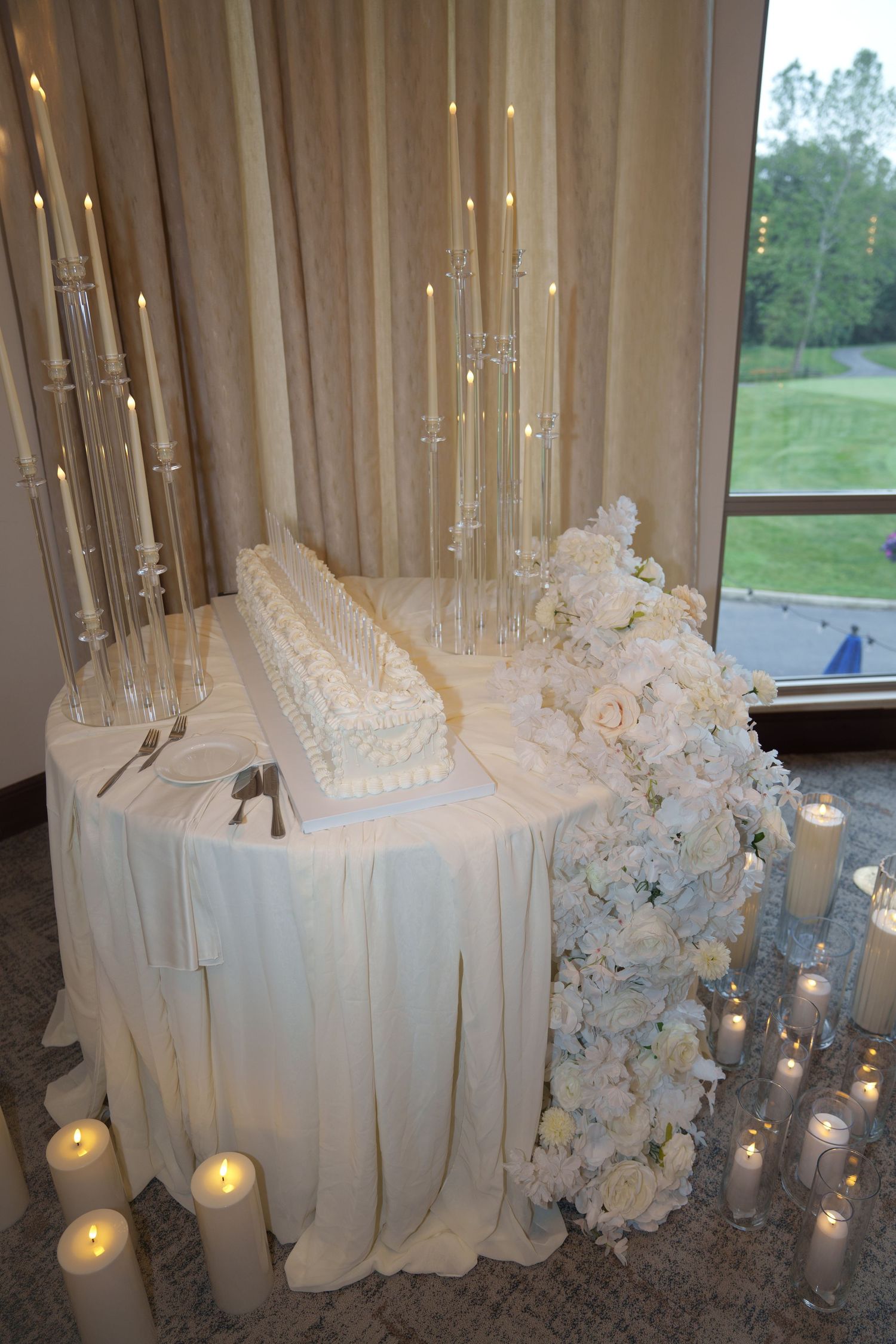 Elegant white wedding cake table with crystal candelabras, white roses, and pillar candles by window.