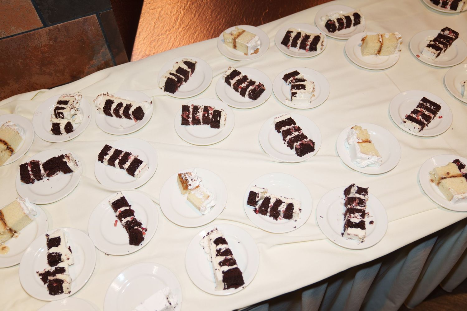 Rows of white plates with chocolate and vanilla cake slices neatly arranged on a white tablecloth at a catered event.