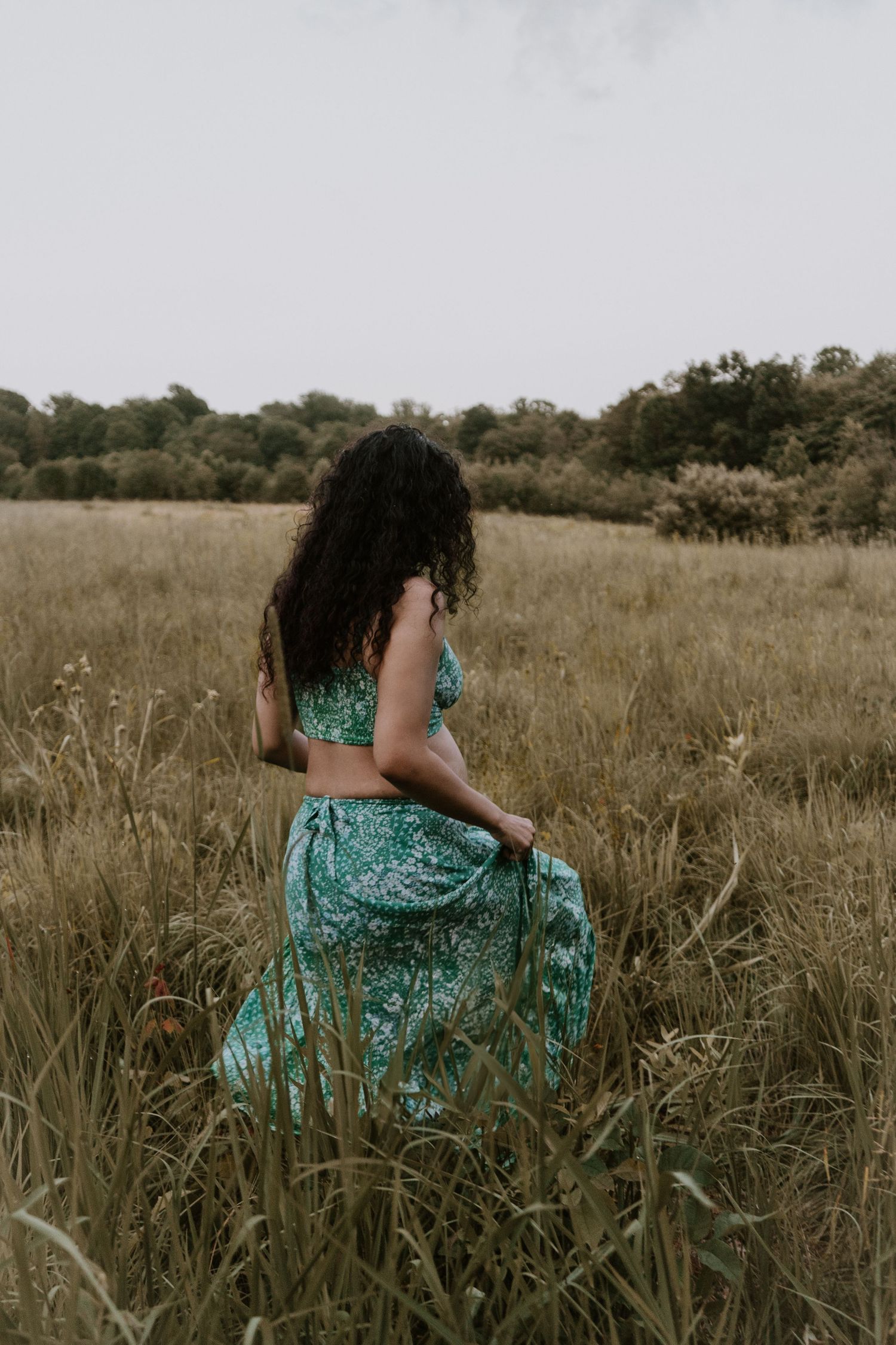 A person in a turquoise patterned dress stands facing away in a golden field of tall grass at sunset.