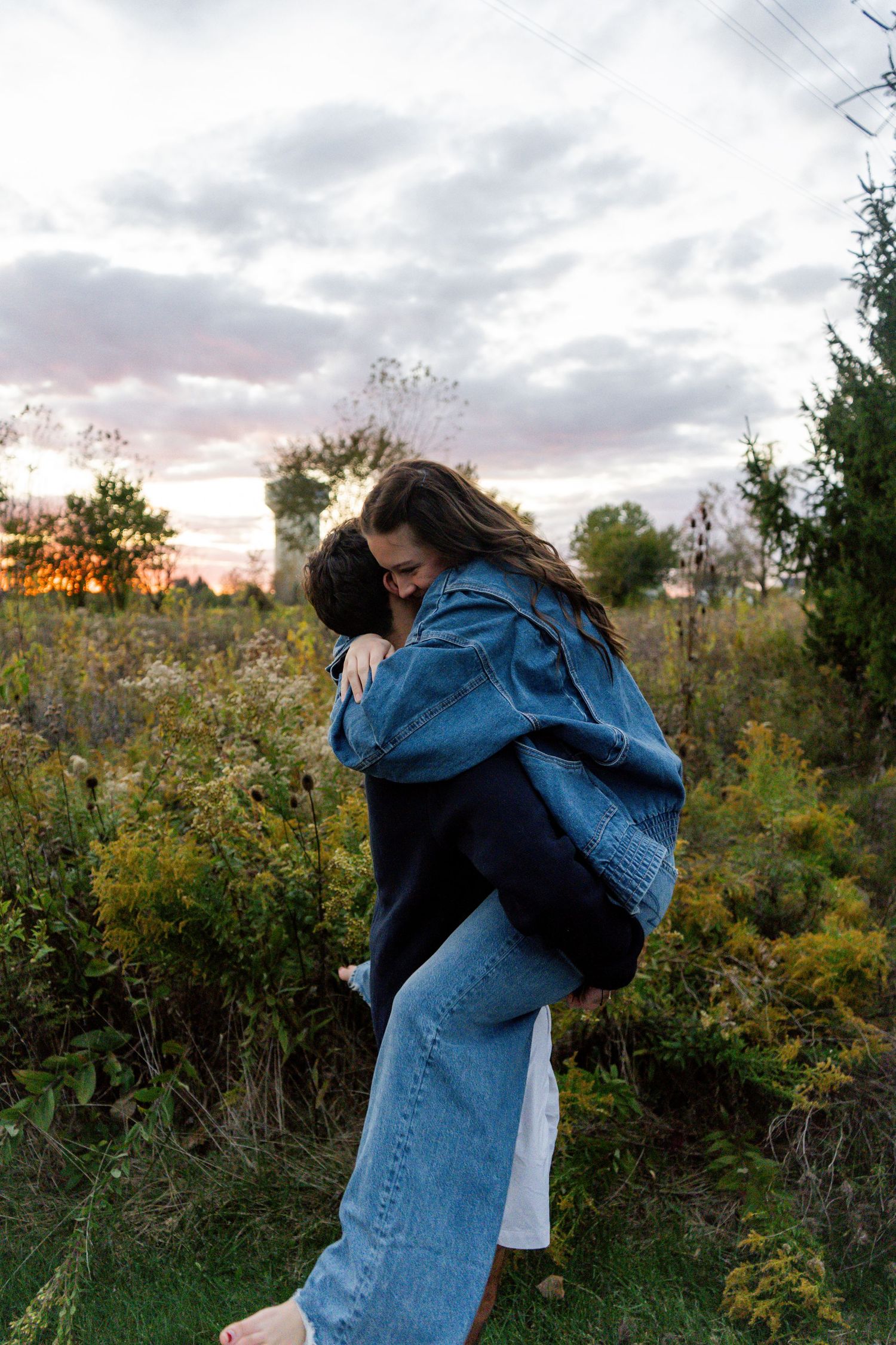 Two people embrace in a warm hug against an autumn sunset backdrop with trees and cloudy skies.