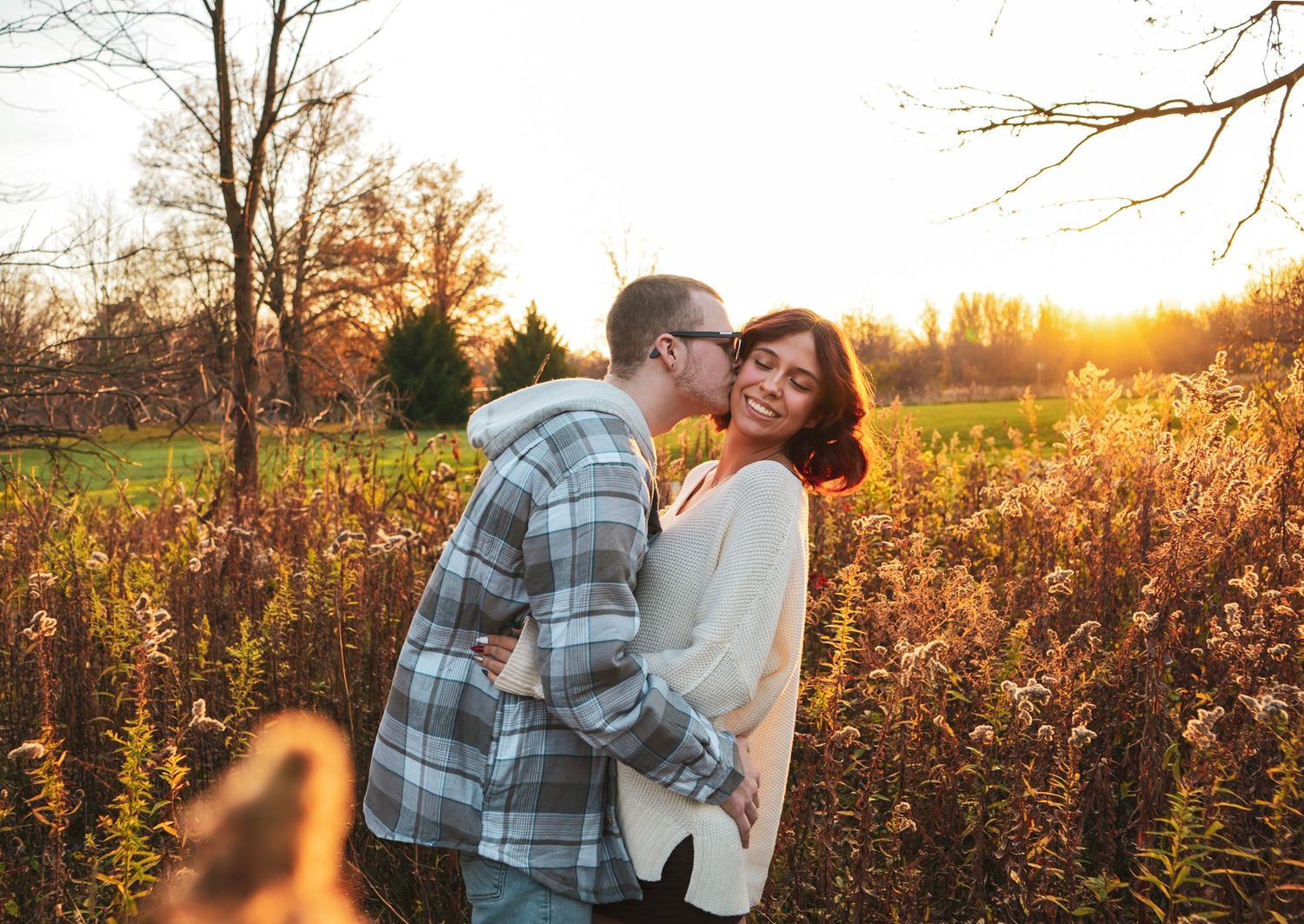 A couple embraces in a field during golden hour with autumn foliage in the background.