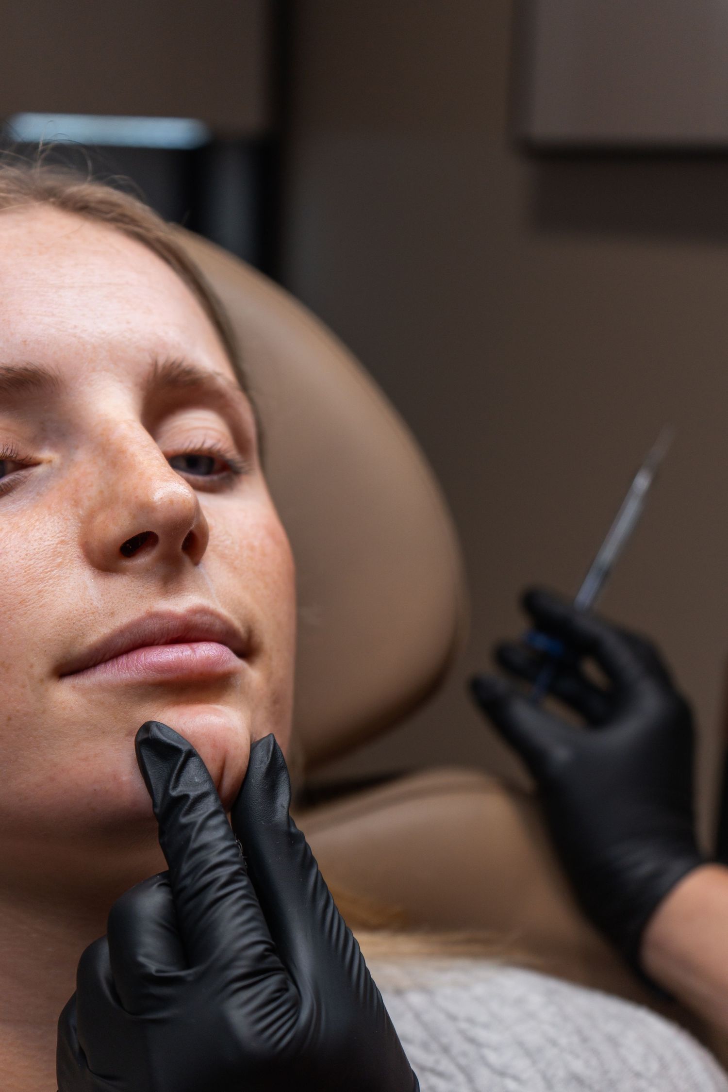 Medical professional wearing black gloves examines patient in dental chair during cosmetic procedure.
