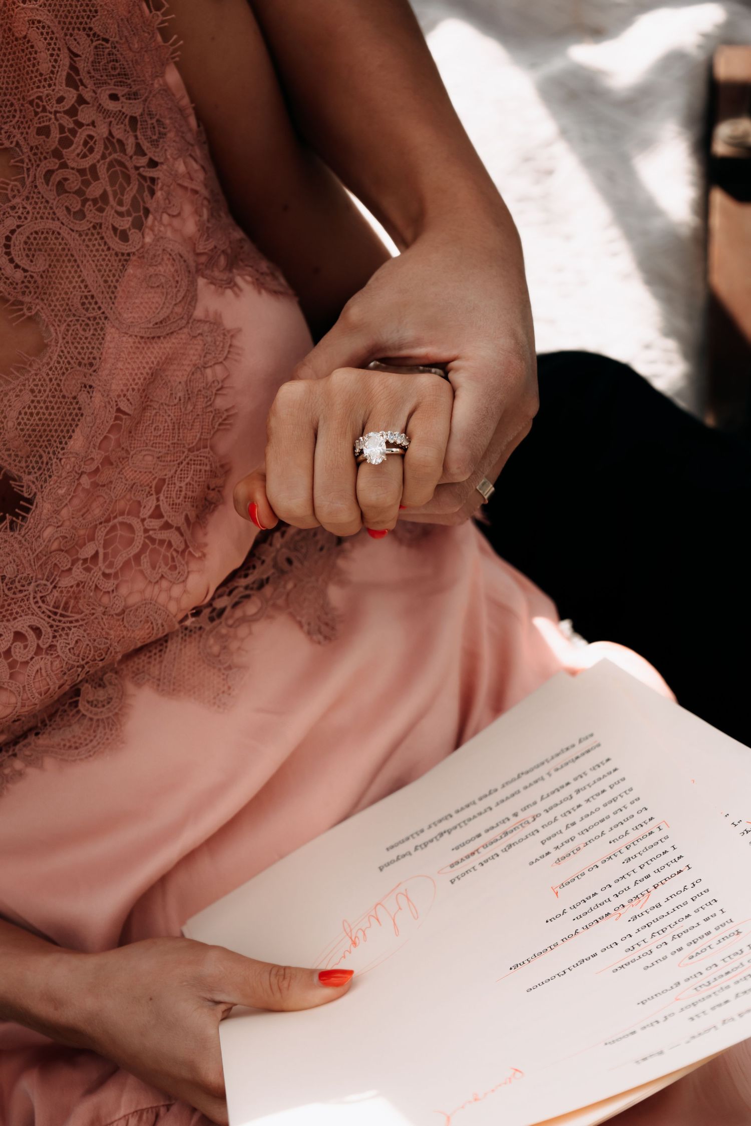 Close-up of a woman in a pink lace dress wearing a diamond ring, holding an open book with orange text.