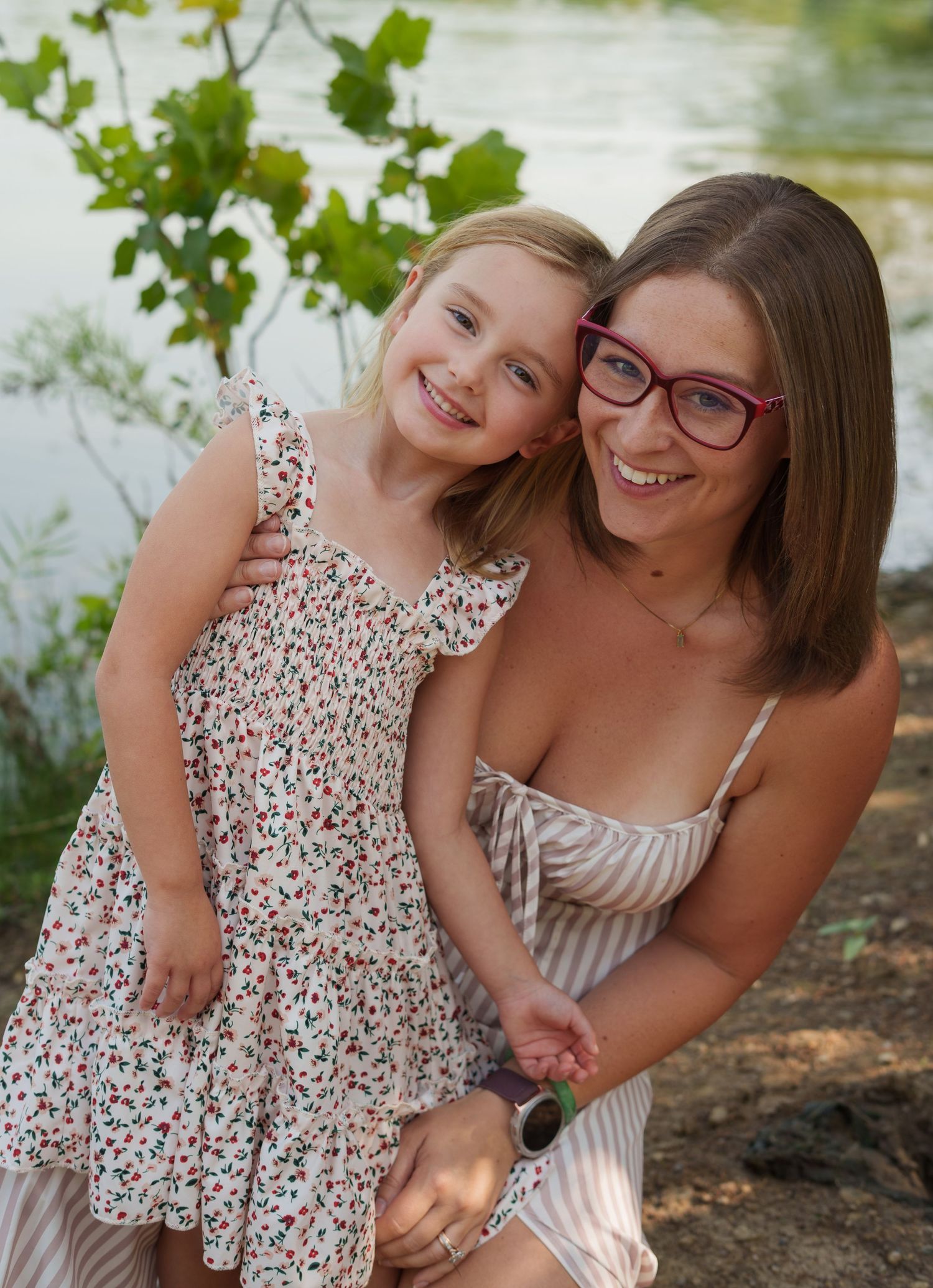 A smiling woman with red glasses hugs a young girl in a floral dress outdoors near a lake.