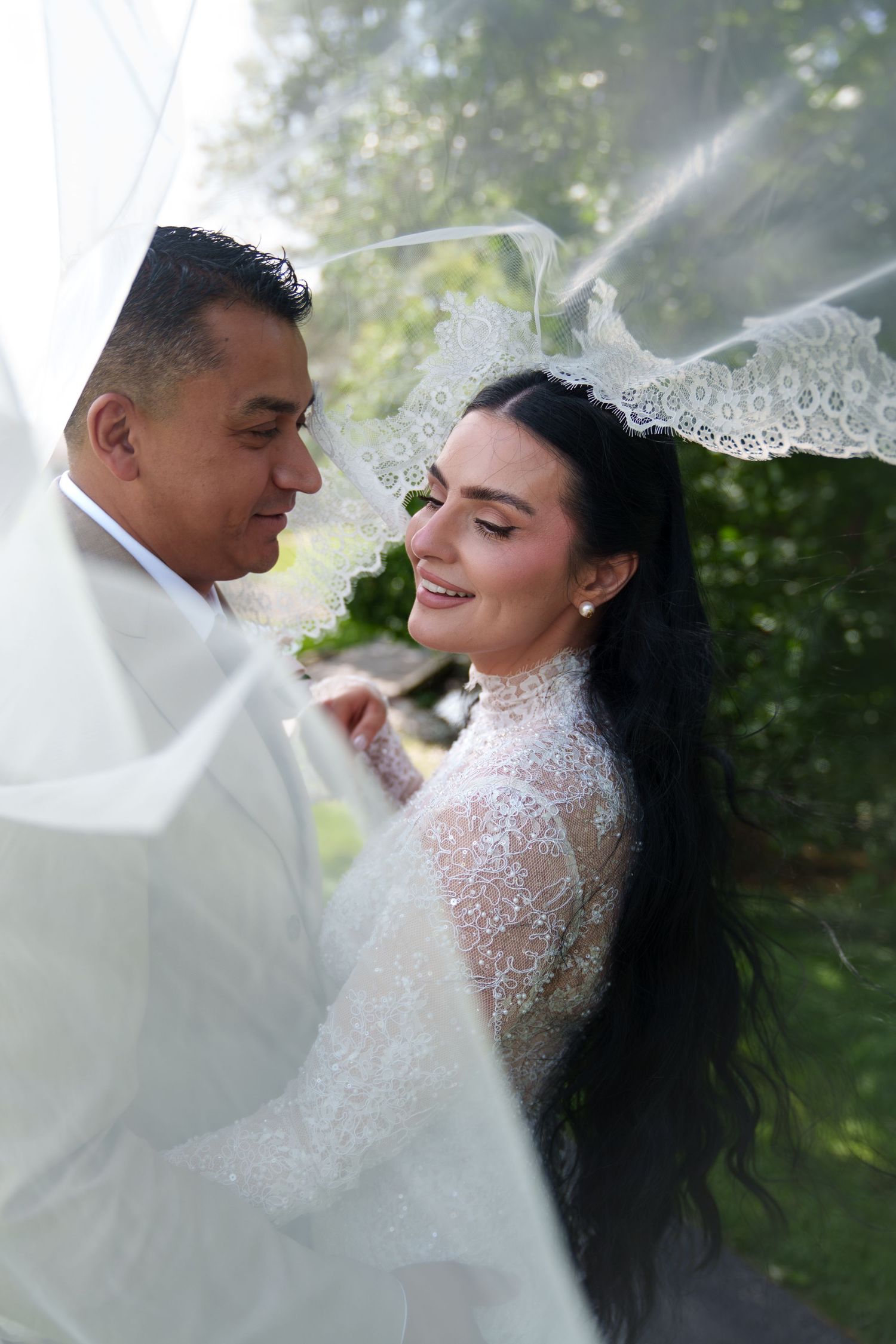 A bride in lace gown and veil smiles at her groom outdoors in a romantic wedding portrait.