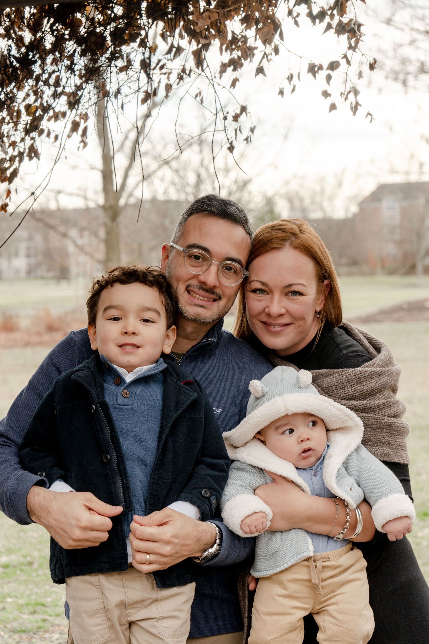 A family of four poses together outdoors on a fall day with trees and buildings in the background.