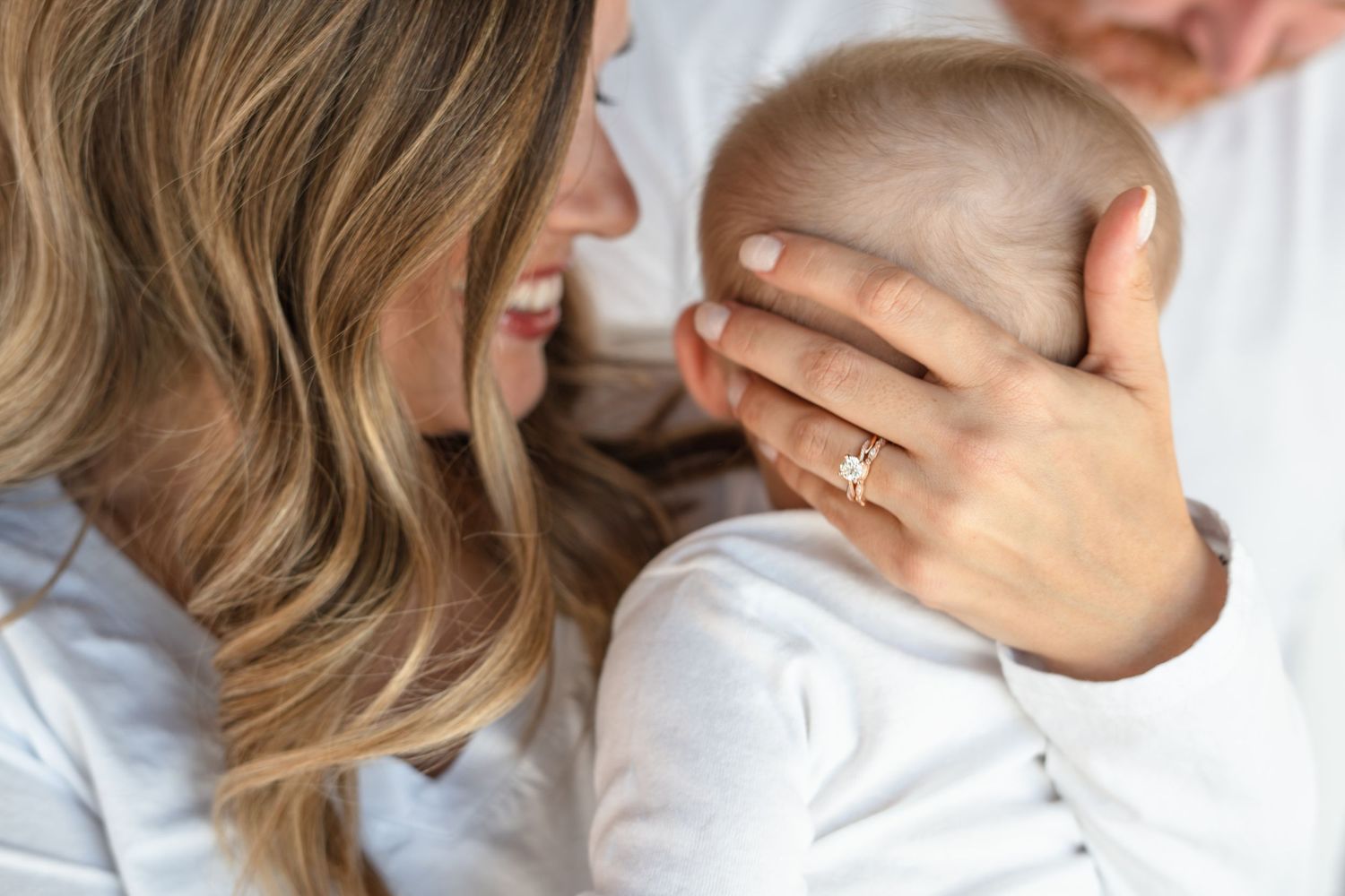 A smiling mother with blonde hair holds her newborn baby while wearing a gold diamond engagement ring.