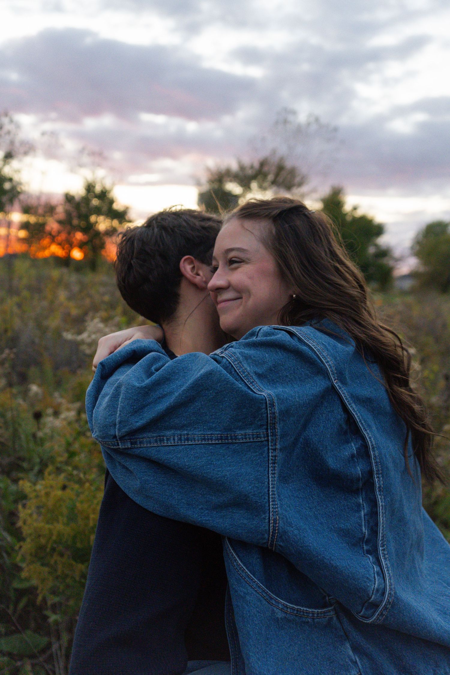 Two people embrace warmly outdoors during golden hour with autumn foliage and cloudy skies behind them.