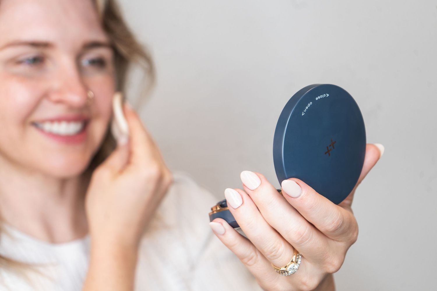 Someone holds a small round compact mirror while talking on a mobile phone in a bright indoor setting.