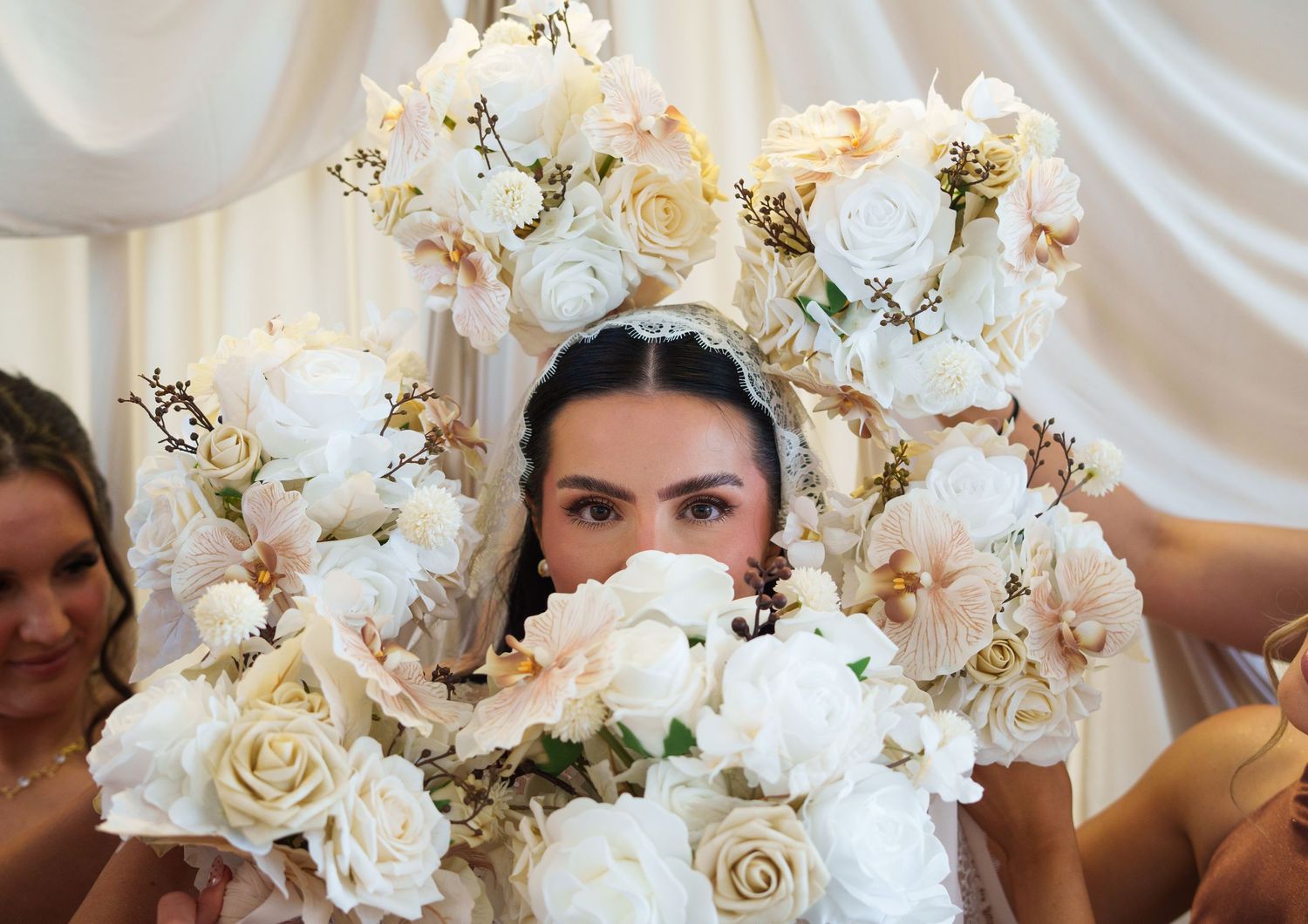 Bride surrounded by white roses and floral headpiece, peering over bouquet at wedding ceremony.