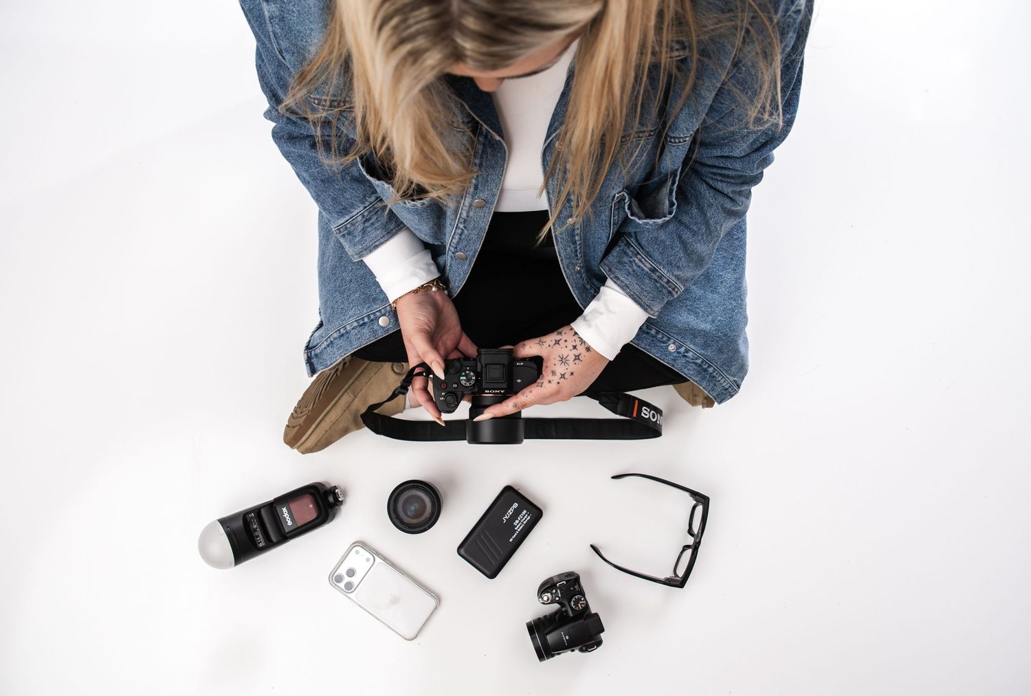 Overhead view of a woman in a denim jacket examining camera gear and accessories on a white surface.
