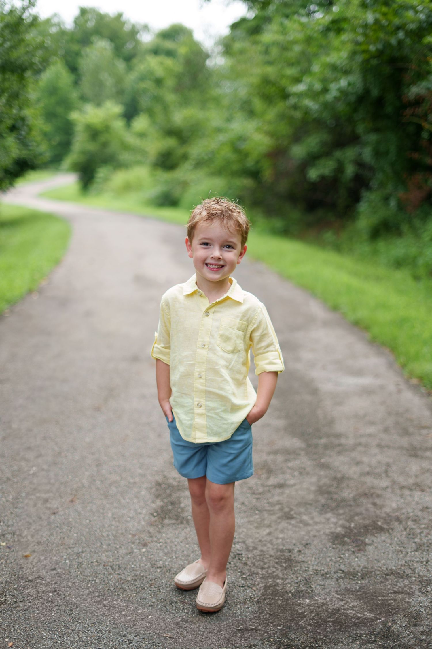 Smiling young boy in yellow shirt and blue shorts standing on a tree-lined path outdoors.