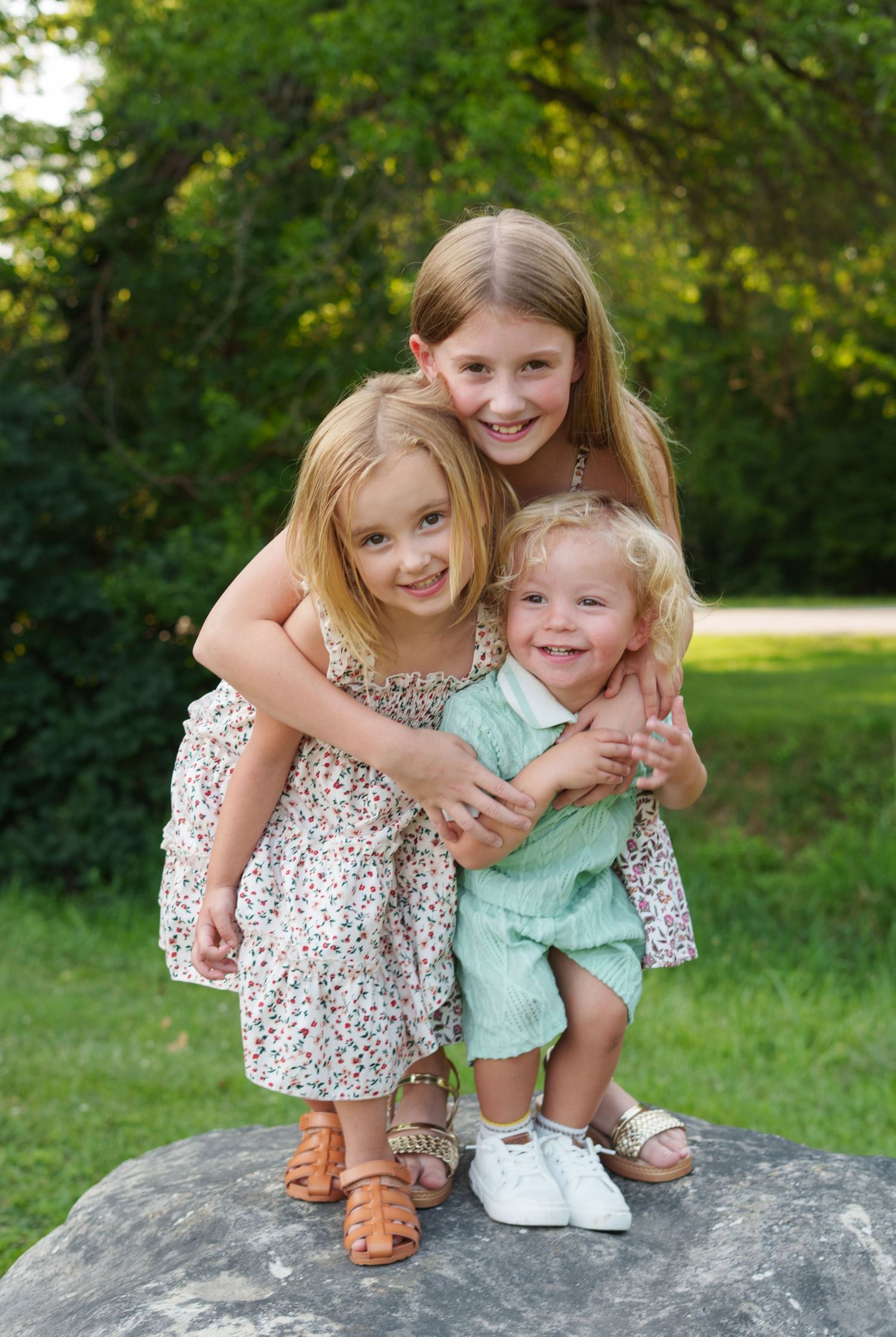 Three blonde siblings pose on a rock outdoors, two girls in floral dresses and a toddler boy in mint green romper.