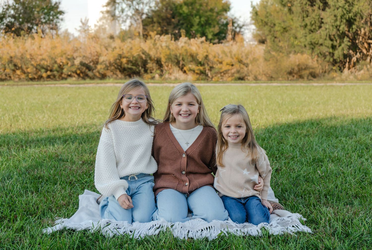 Three smiling girls sitting together on a blanket in a grassy field during autumn, wearing cozy neutral-toned sweaters.