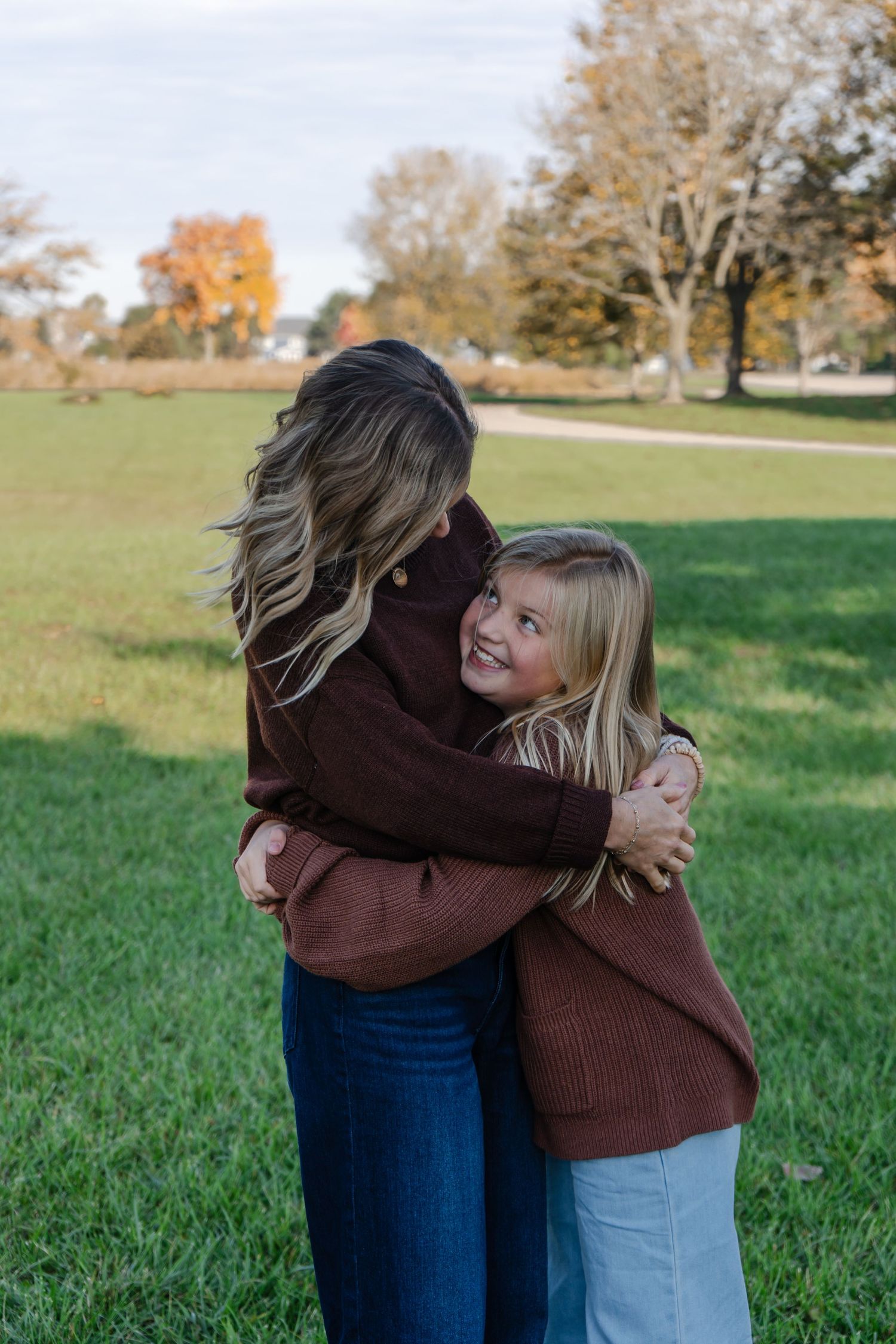 Two people share a warm embrace in a grassy field on an autumn day with trees in the background.