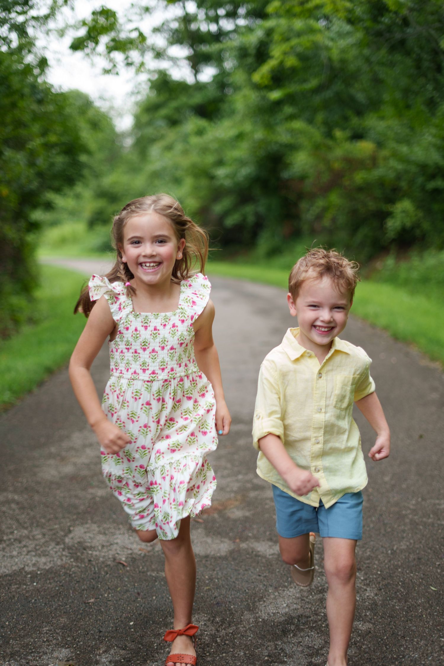Two happy children running on a tree-lined path, girl in floral dress and boy in yellow shirt and blue shorts.