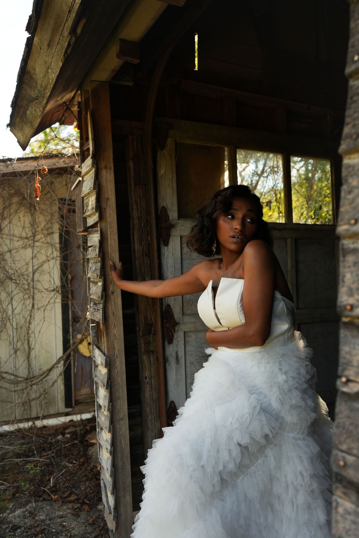 A person in a white ruffled wedding gown poses against rustic wooden walls in an abandoned building.