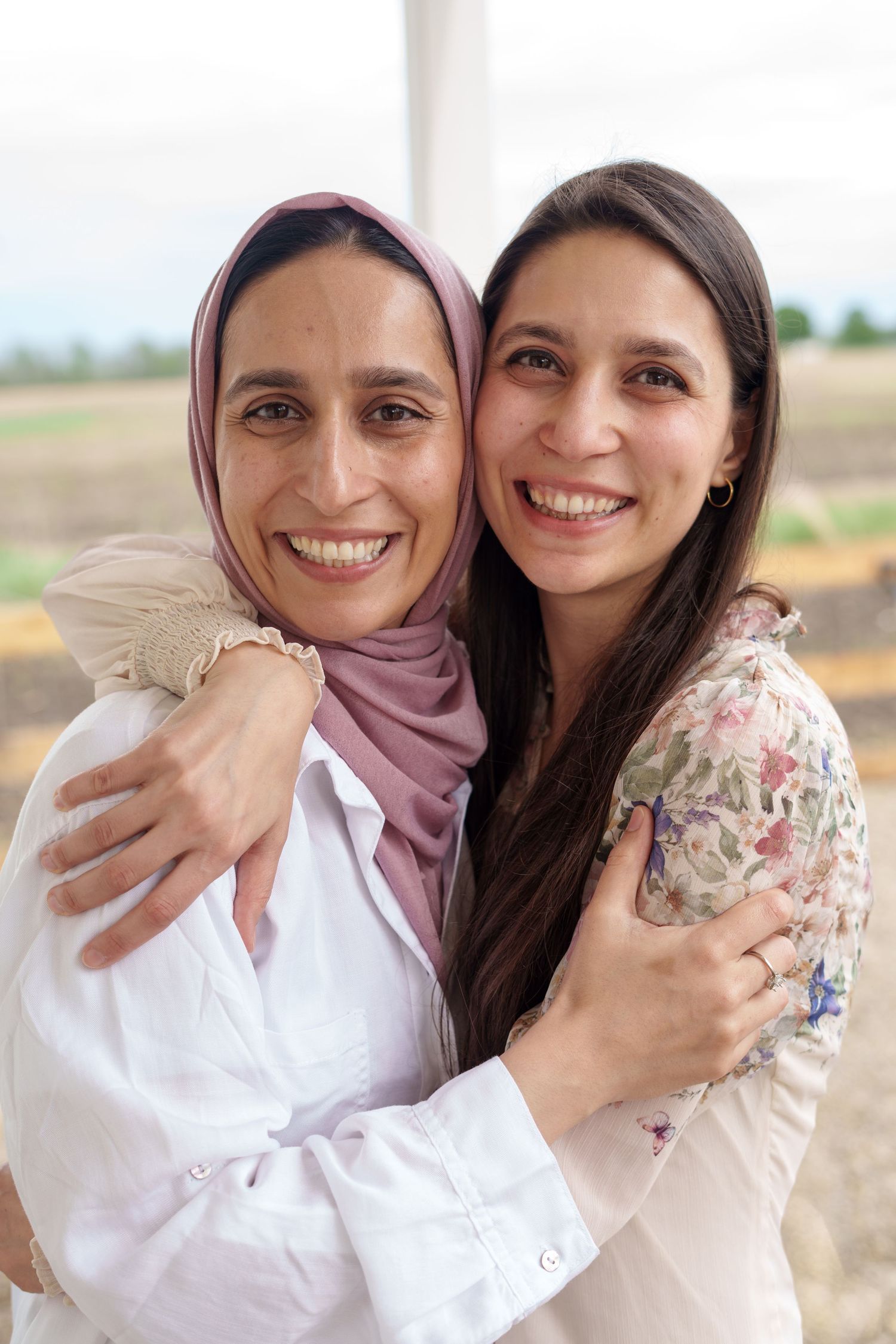 Two smiling women hugging outdoors, one wearing a pink hijab and white shirt, the other in a floral blouse.