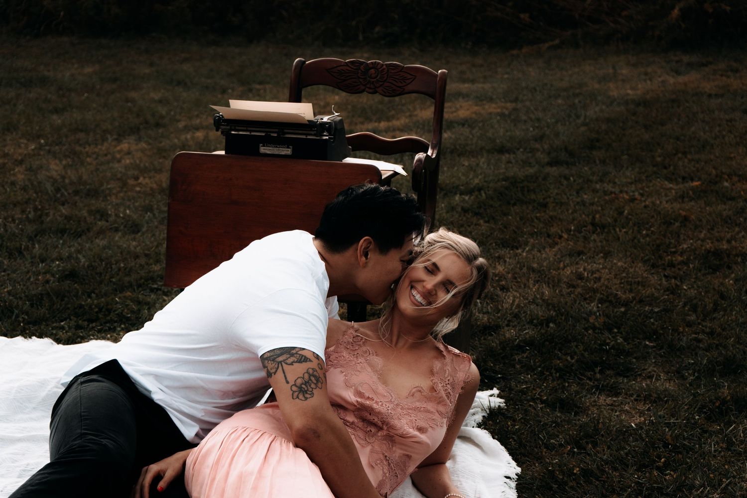 Couple sharing a romantic moment on a blanket in a grassy field with a vintage typewriter nearby.