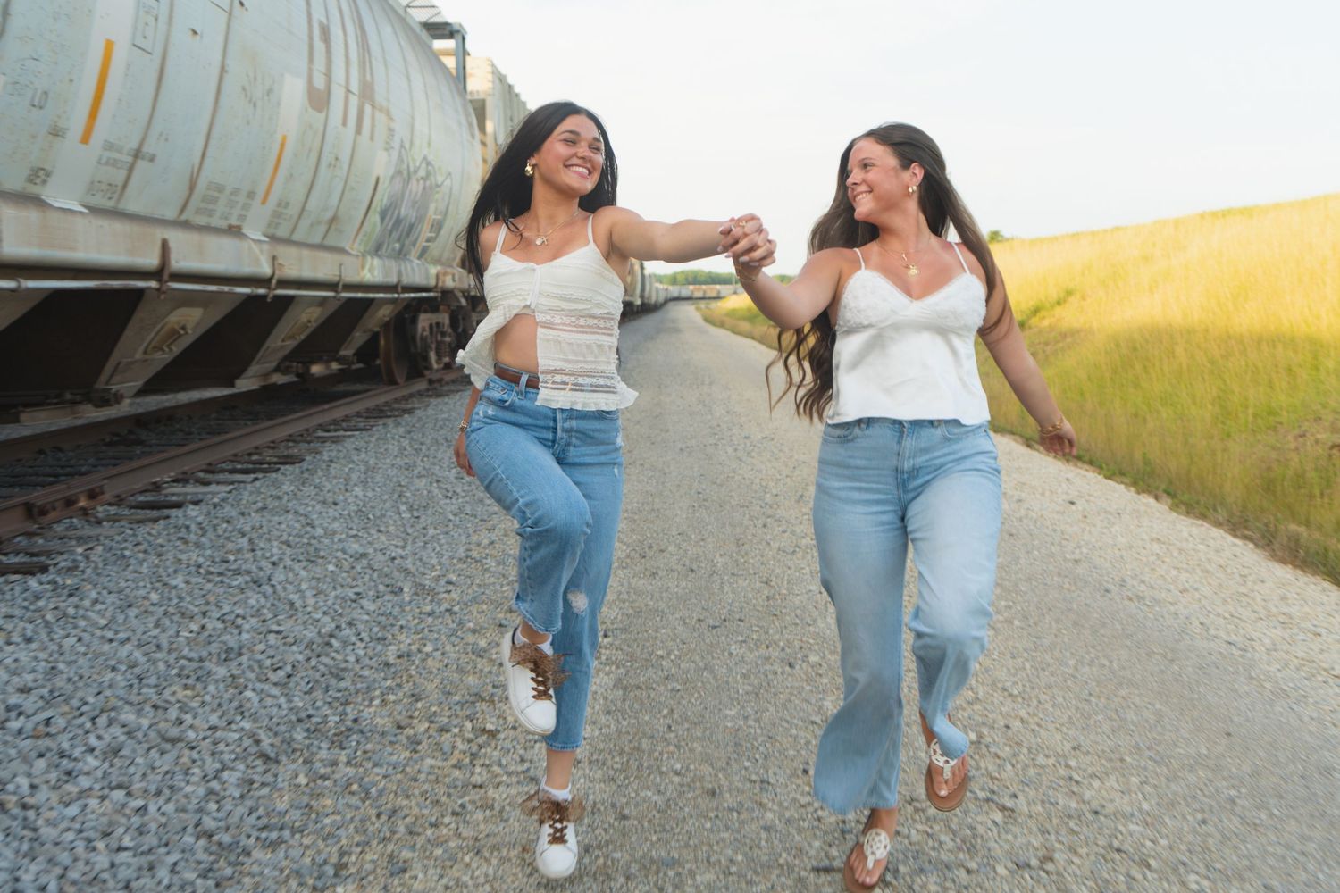 Two friends in casual summer outfits running joyfully along a railroad track next to a golden field at sunset.