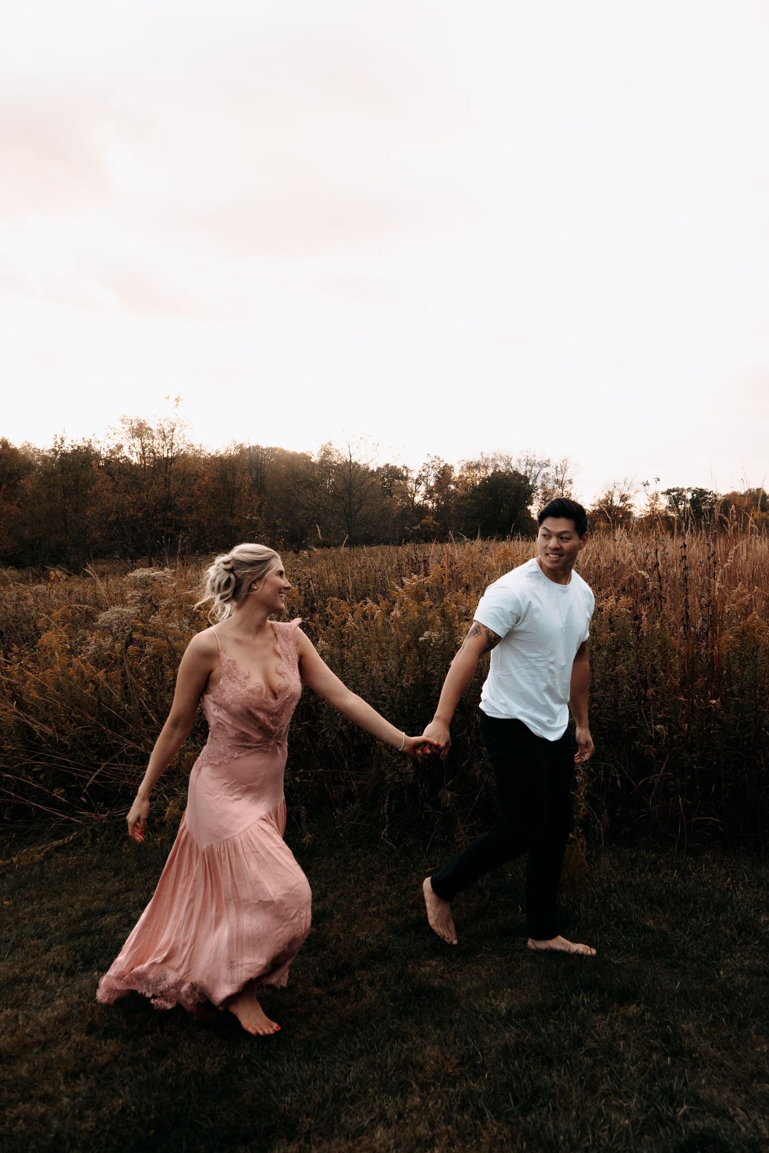 A couple in formal attire dances playfully in a field at sunset creating a romantic silhouette against the sky.