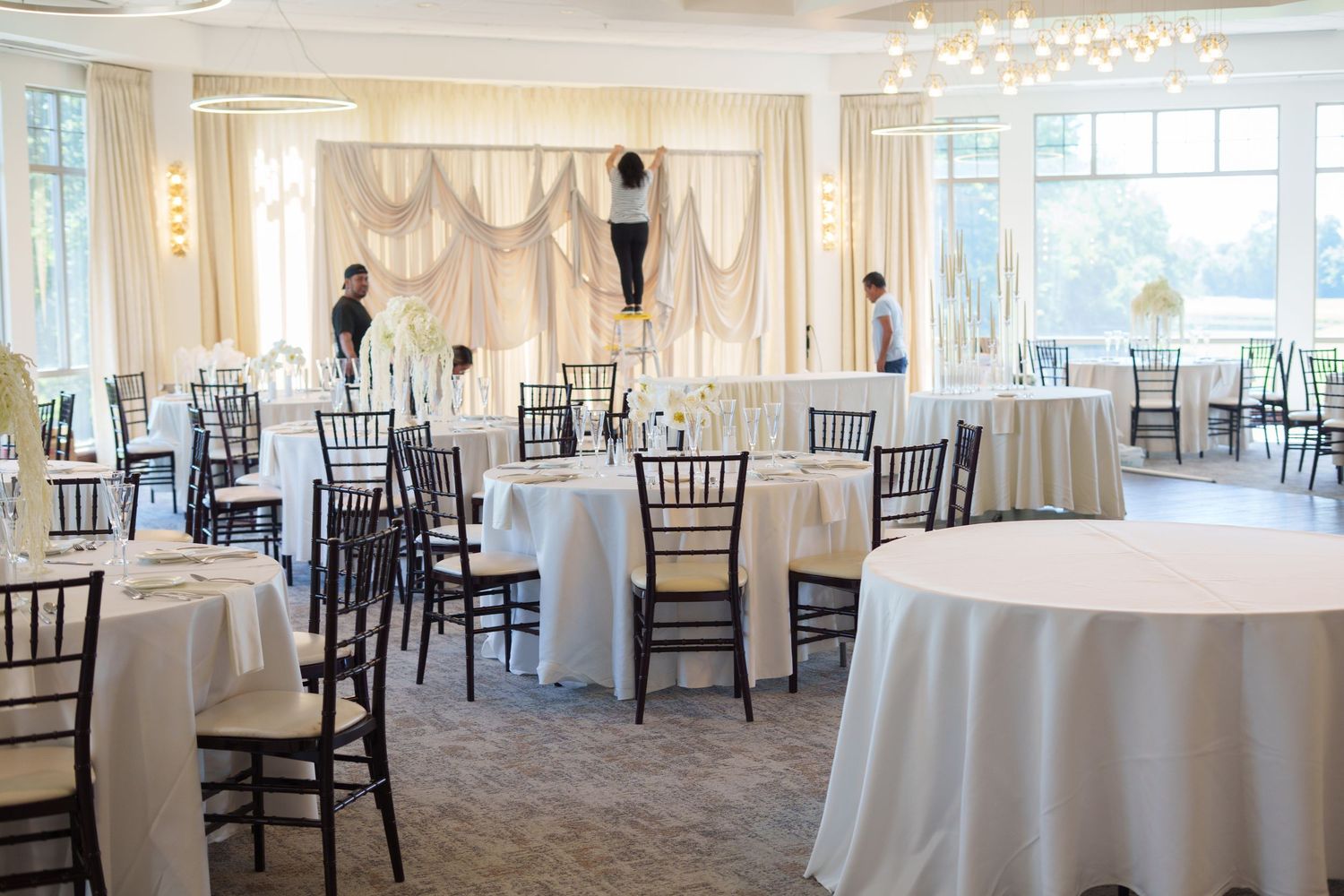 Elegant wedding reception venue with white draped backdrop, round tables with white linens, and dark Chiavari chairs in a bright ballroom.
