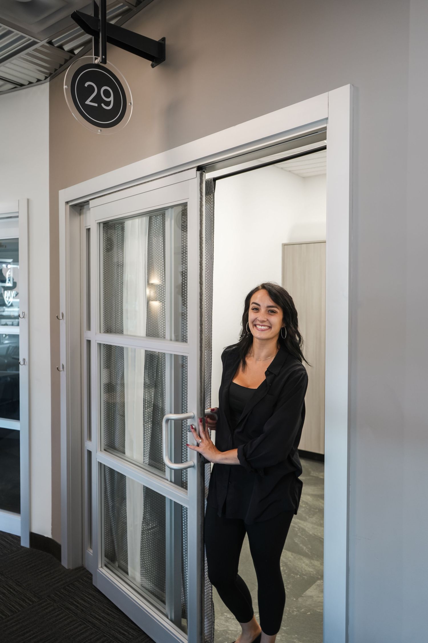 A person in black clothing poses playfully in a modern office hallway with white walls and numbered doors.