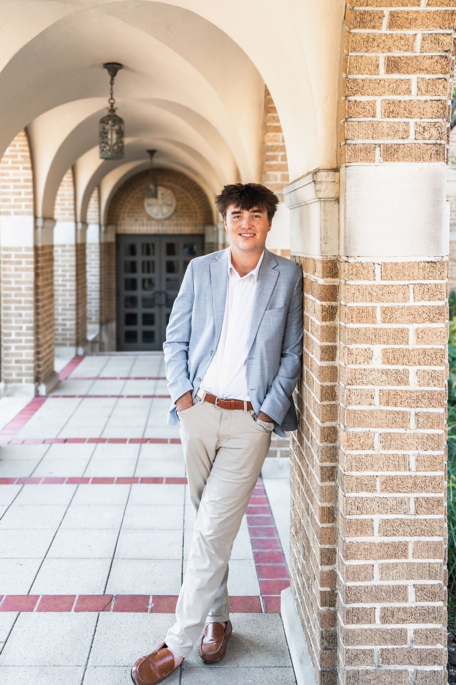 Person in light colored suit leans casually against brick archway in outdoor covered walkway.