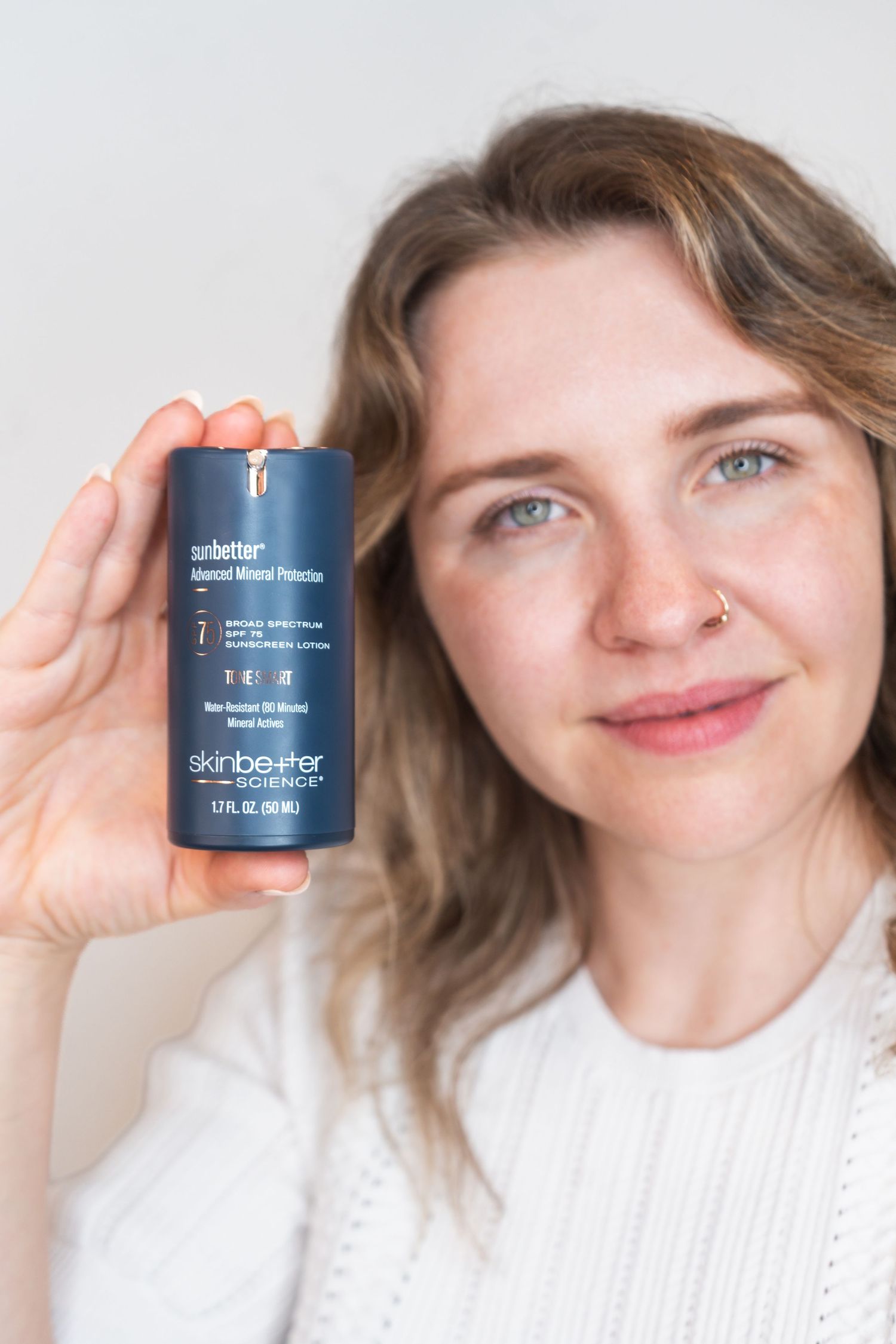 Someone holding up a navy blue skincare product container while wearing a white knit sweater and smiling at the camera.