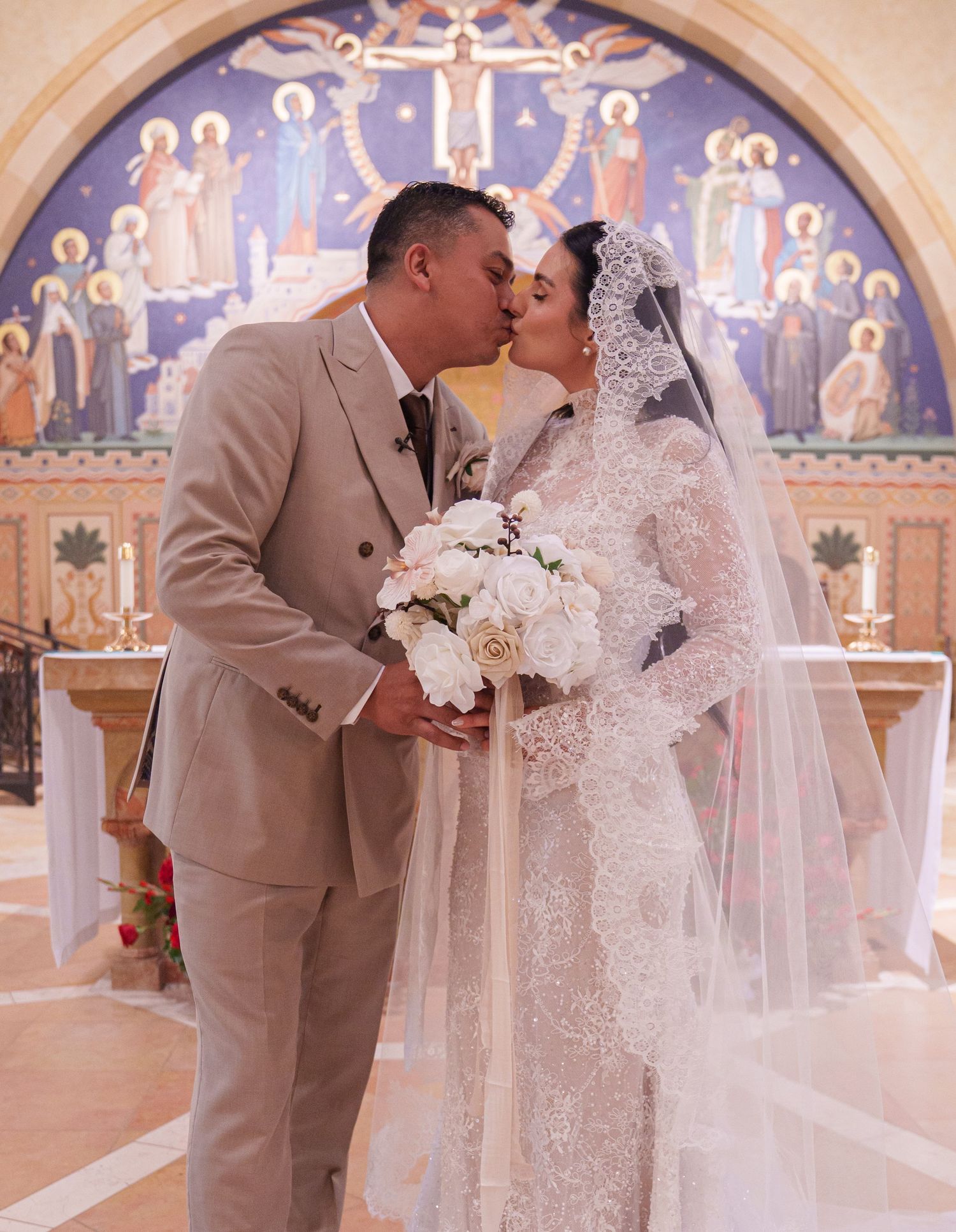Bride and groom share a kiss at the altar of a decorated church, bride wearing lace gown and veil holding white bouquet.
