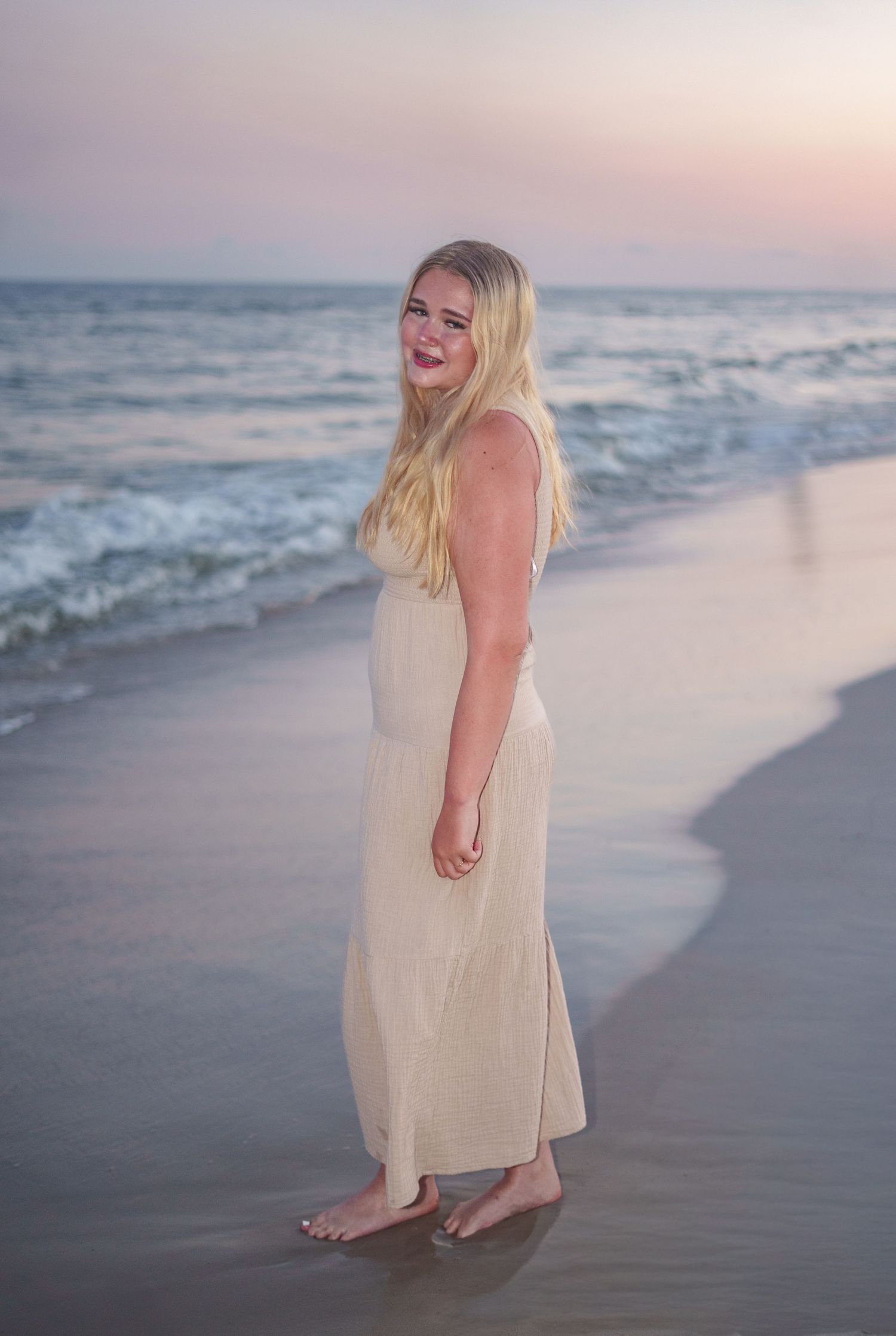 Blonde woman in a cream maxi dress standing barefoot on a sandy beach at sunset with gentle waves.