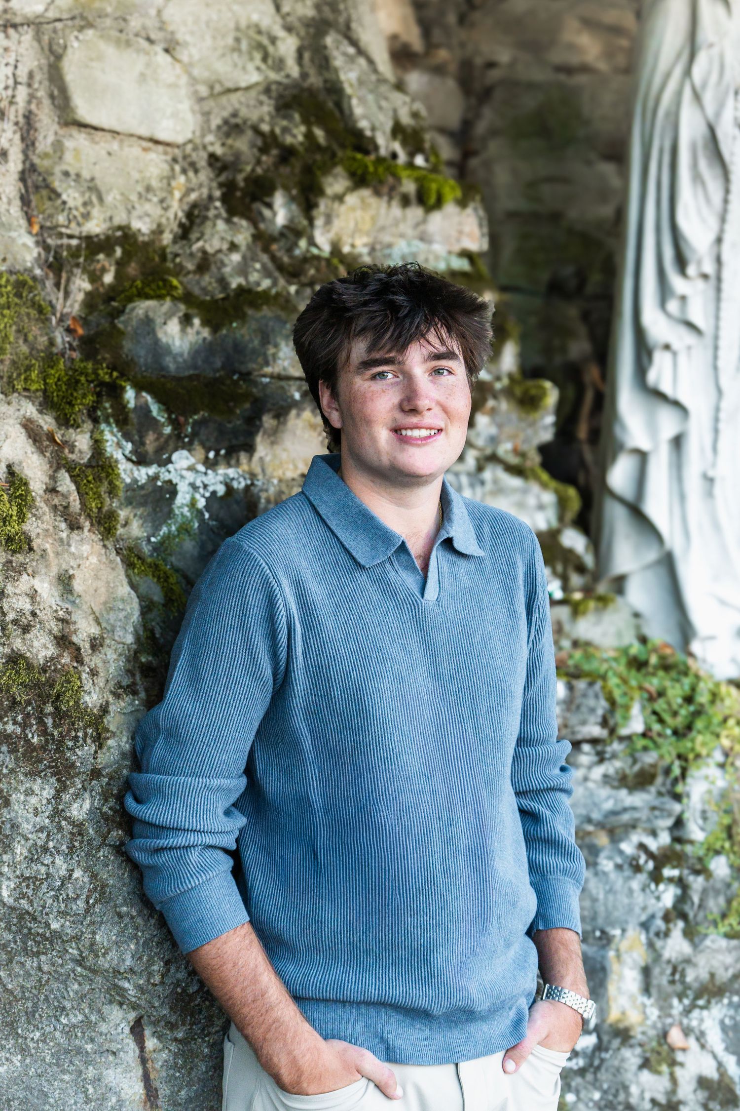 A person in a blue sweater stands against a mossy stone wall with a relaxed pose and casual style.
