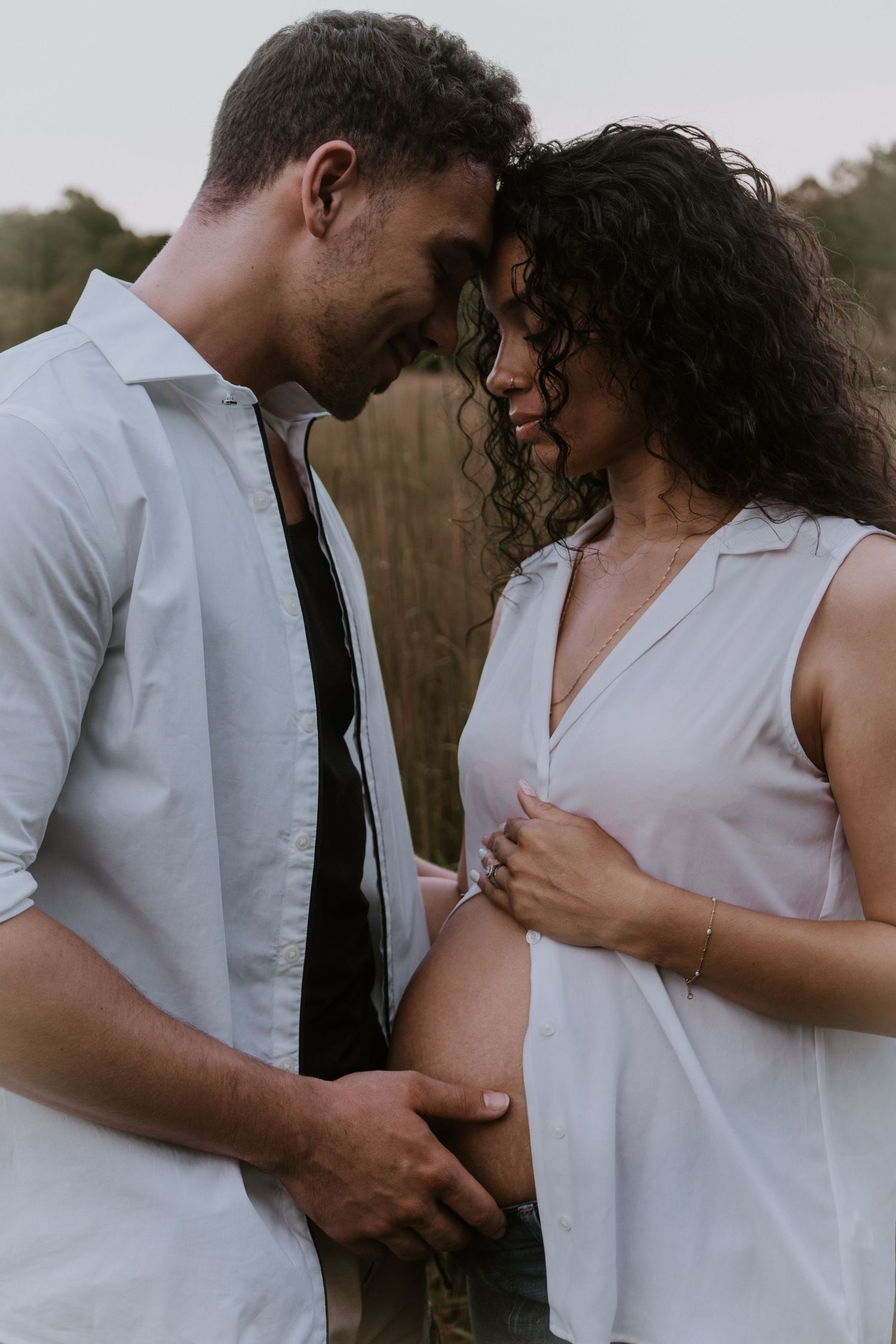 Intimate maternity photo of couple in white outfits sharing a tender moment in a natural outdoor setting at dusk.
