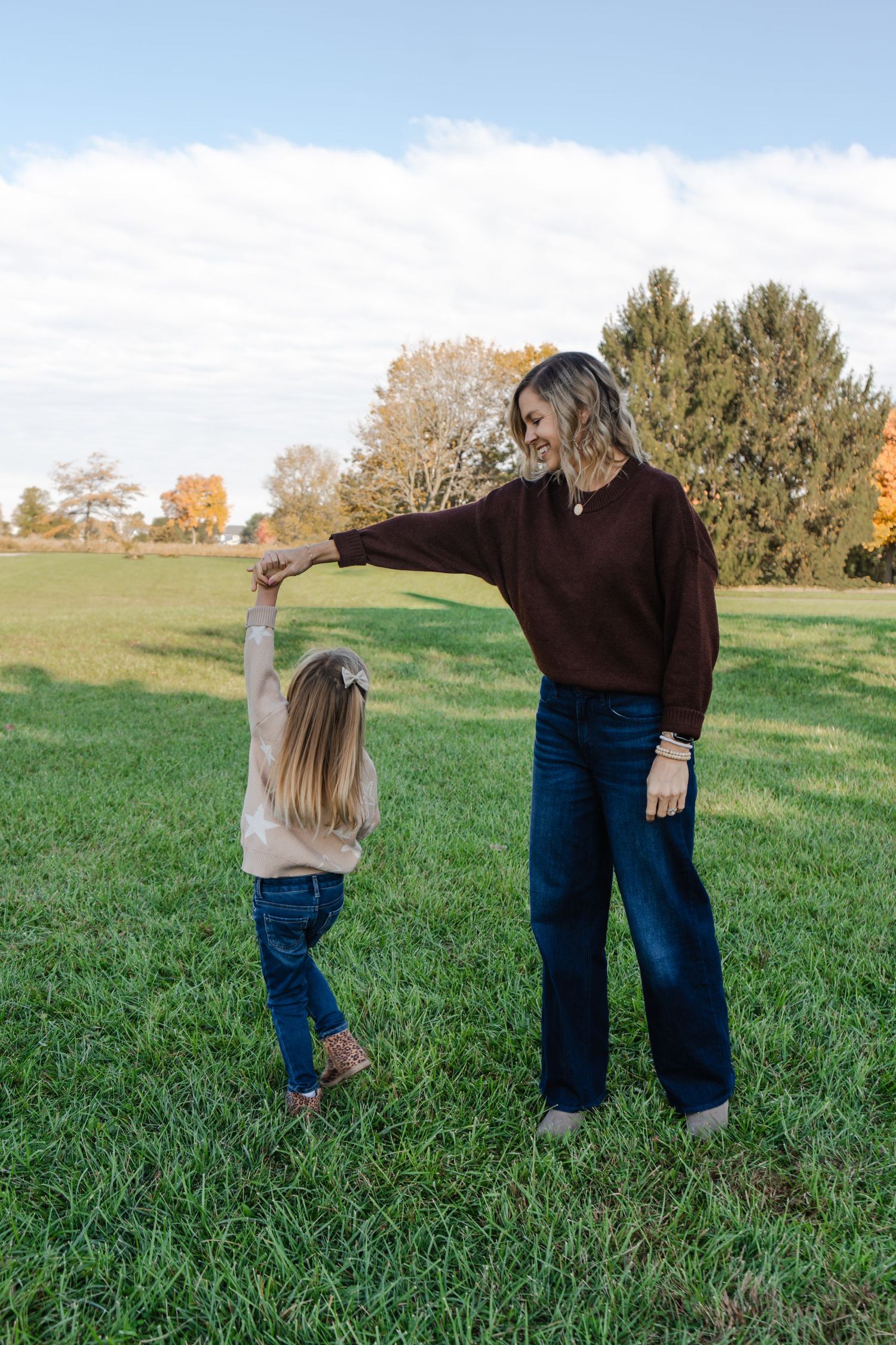 Two figures in matching brown sweaters and jeans hold hands while standing together in a grassy field on an autumn day.
