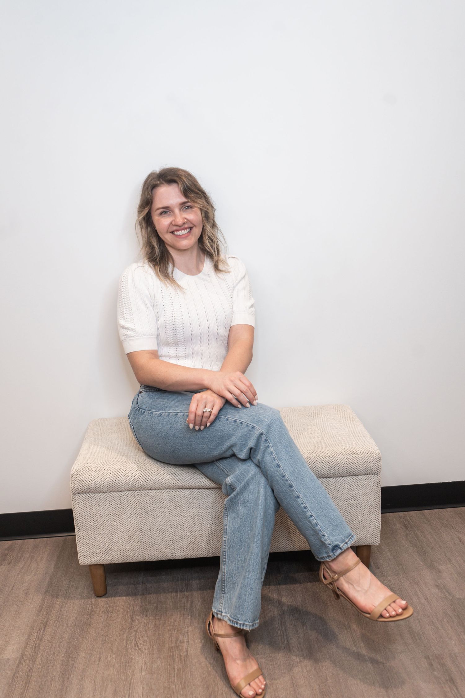 Someone wearing a white t-shirt and blue jeans sits casually on a beige bench with tan sandals.