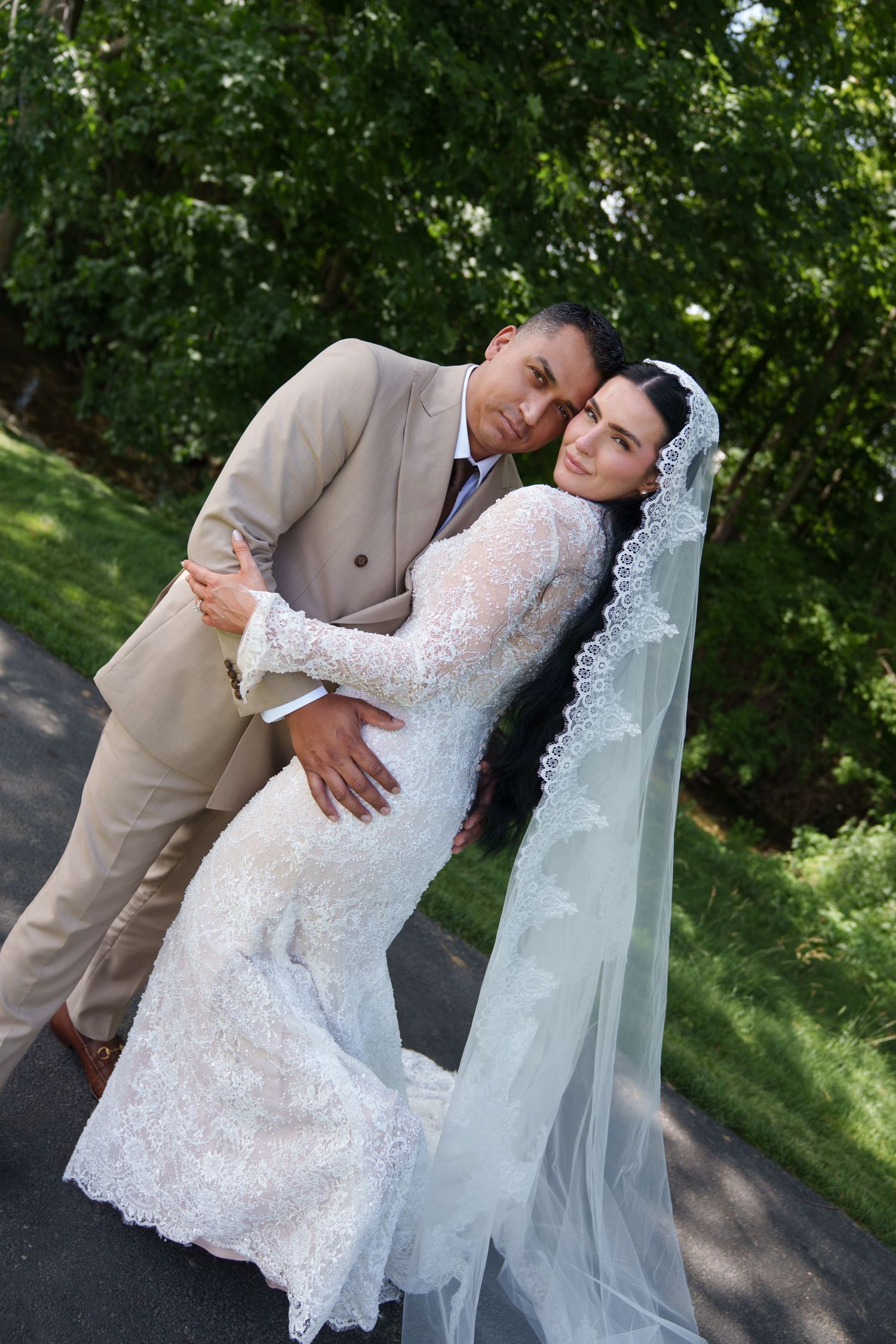 Bride in lace gown and veil embraces groom in tan suit outdoors surrounded by lush green trees on their wedding day.