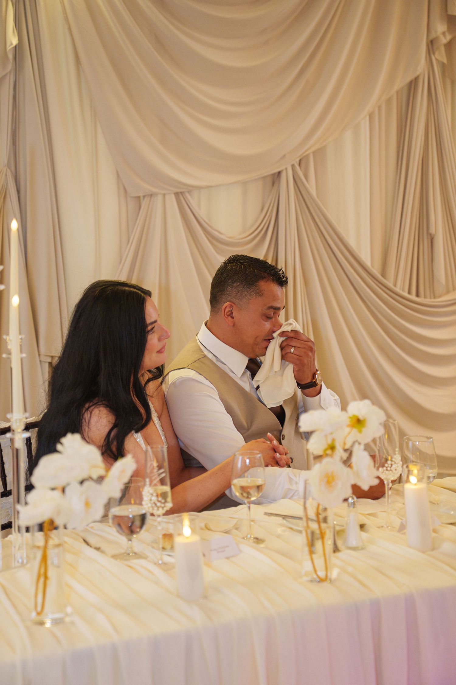 Bride and groom share an emotional moment at their elegant wedding reception table with white drapes and candles.