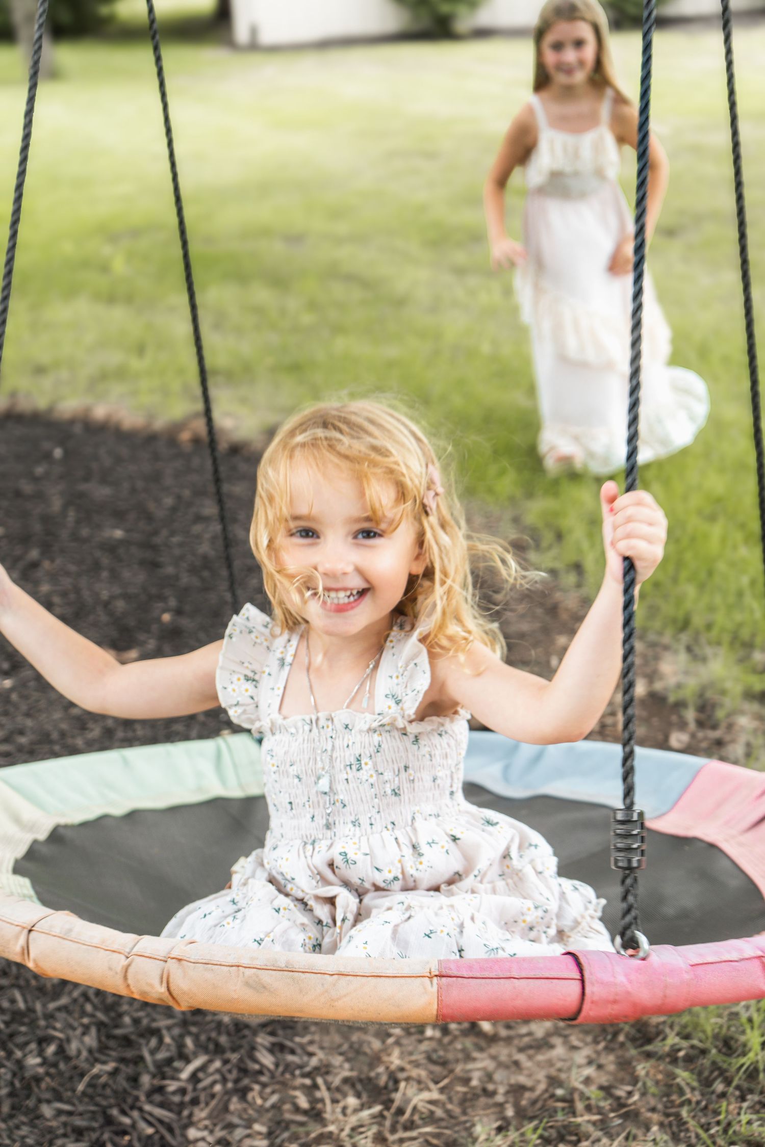 Child in white dress enjoying a round disc swing outdoors while another person stands in background on grass.
