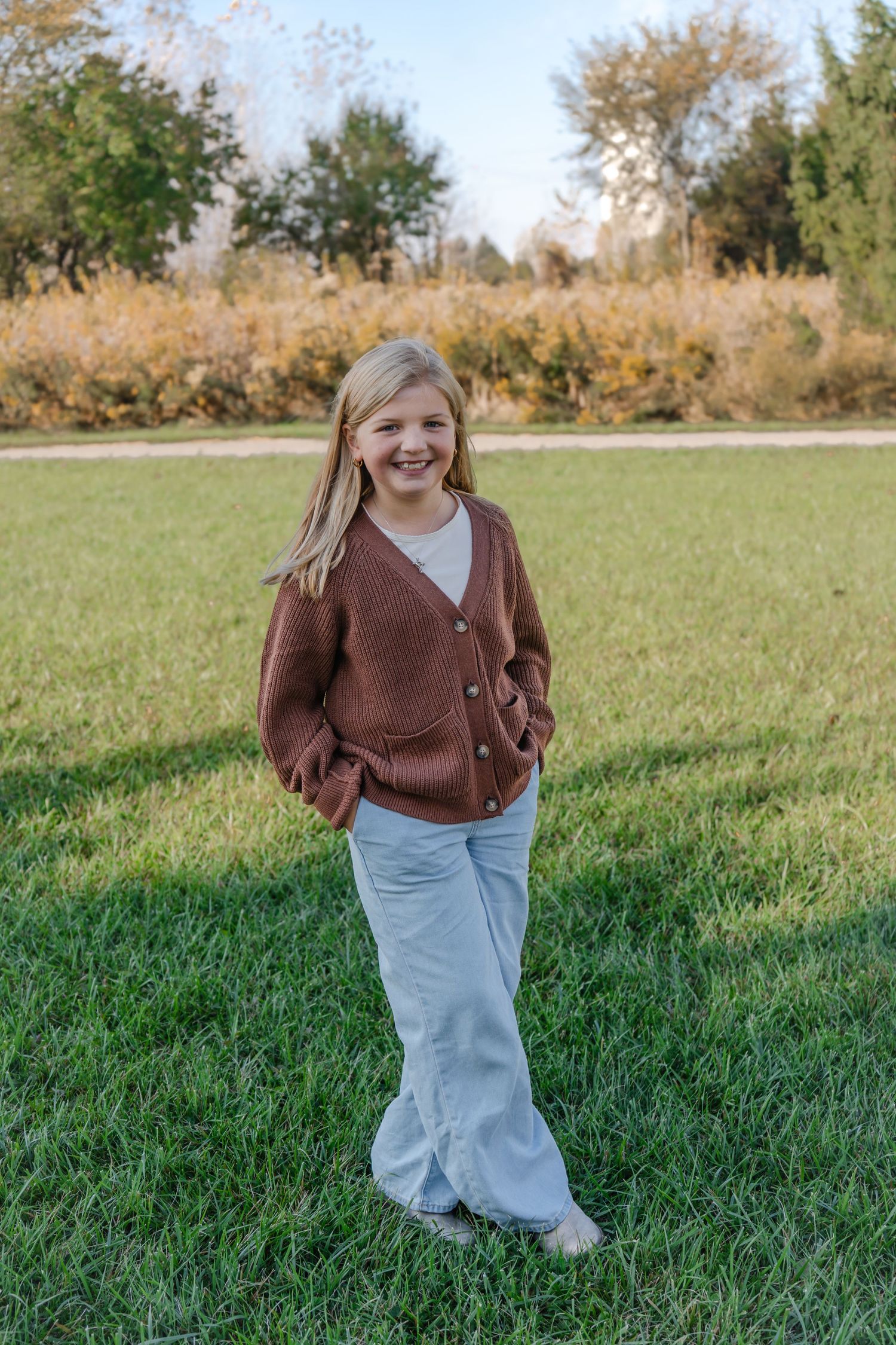 A figure in a brown cardigan and light jeans stands casually on grass with autumn foliage in the background.