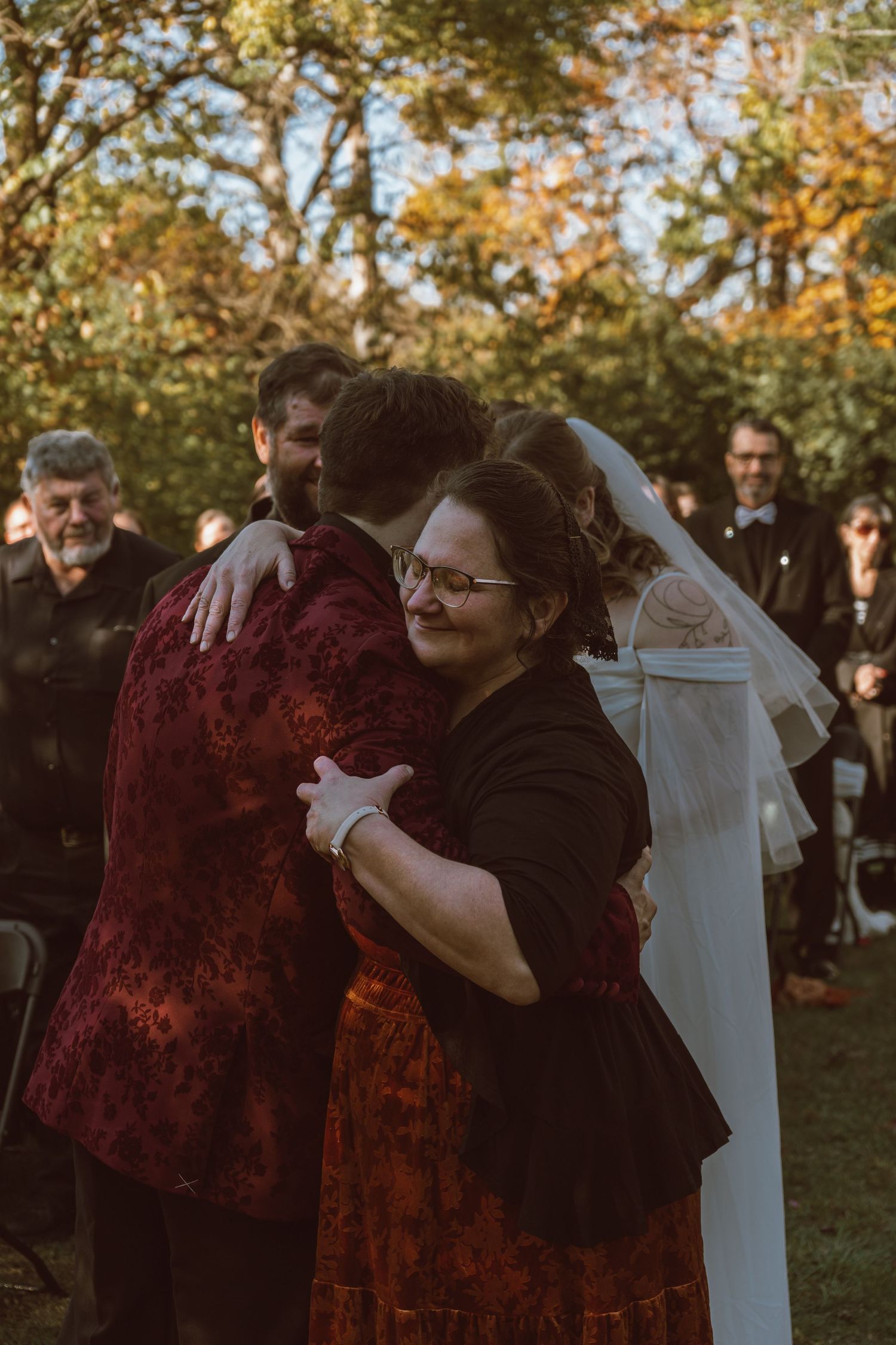 A couple embraces during an outdoor autumn wedding ceremony wearing burgundy and black attire.