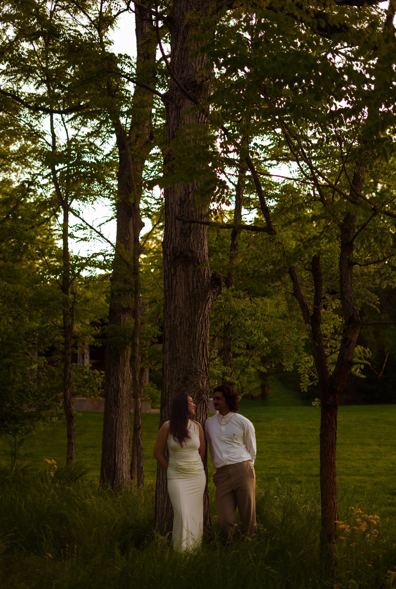 Couple standing together among tall trees in a lush green park during golden hour sunset light.